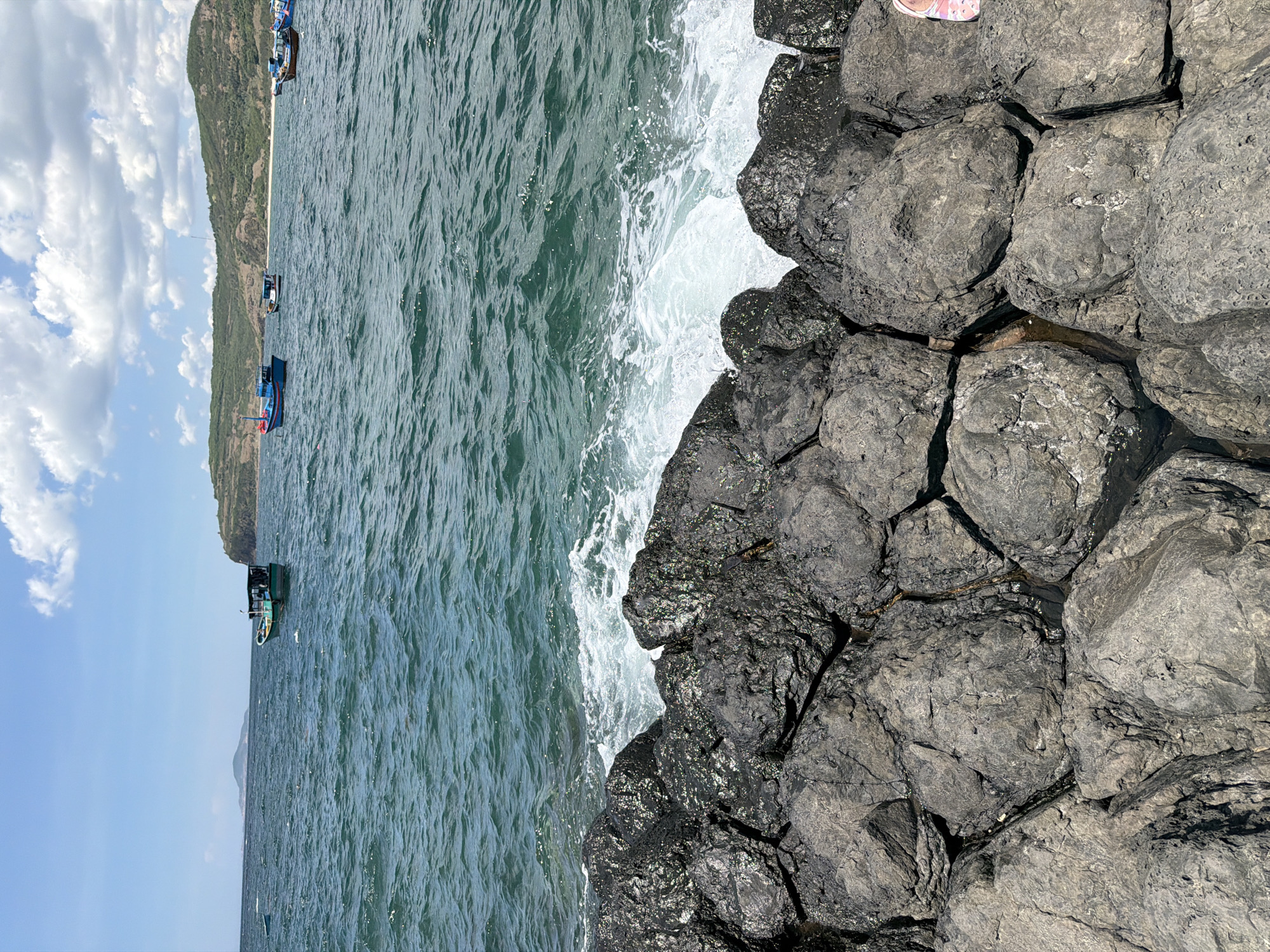 A low-angle, eye-level medium shot captures a rocky shore with the ocean and distant hills under a partly cloudy sky. In the foreground, dark, textured rocks form a natural barrier, with some rocks appearing wet from the waves. The water in the middle ground is a deep blue-green, rippling with white foam as waves crash against the rocks. Several small boats are visible on the water, some closer and some farther away, anchored near the coastline. In the background, green, rolling hills dotted with vegetation rise from the water's edge, creating a natural horizon. The sky is a bright blue with scattered white clouds, suggesting a clear, sunny day. The scene is likely a coastal area in Phú Lương, Vietnam, characterized by its rugged shoreline and seafaring activities. There are no people visible in the immediate foreground or middle ground, but a pair of pink sandals are visible on the far right edge of the frame, suggesting a person is present just outside the primary focus of the image.