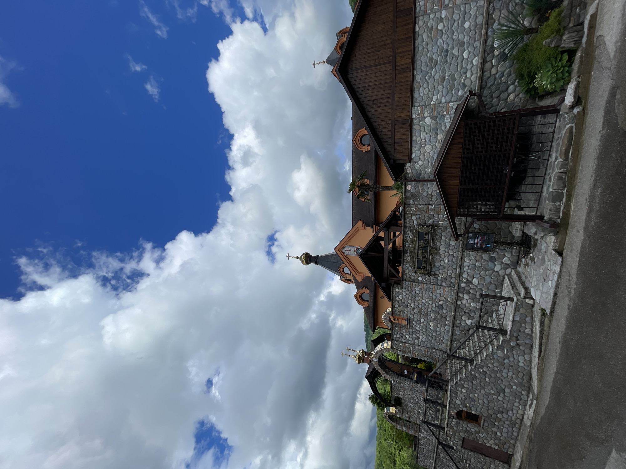 The image shows a partially cloudy sky above the monastery "Cross Empty" in Solokh-Aul, Russia. The architecture of the monastery includes stone walls, wooden roofs, and golden crosses. The building's exterior also features palm trees. A portion of the ground is visible, covered with stone and vegetation.