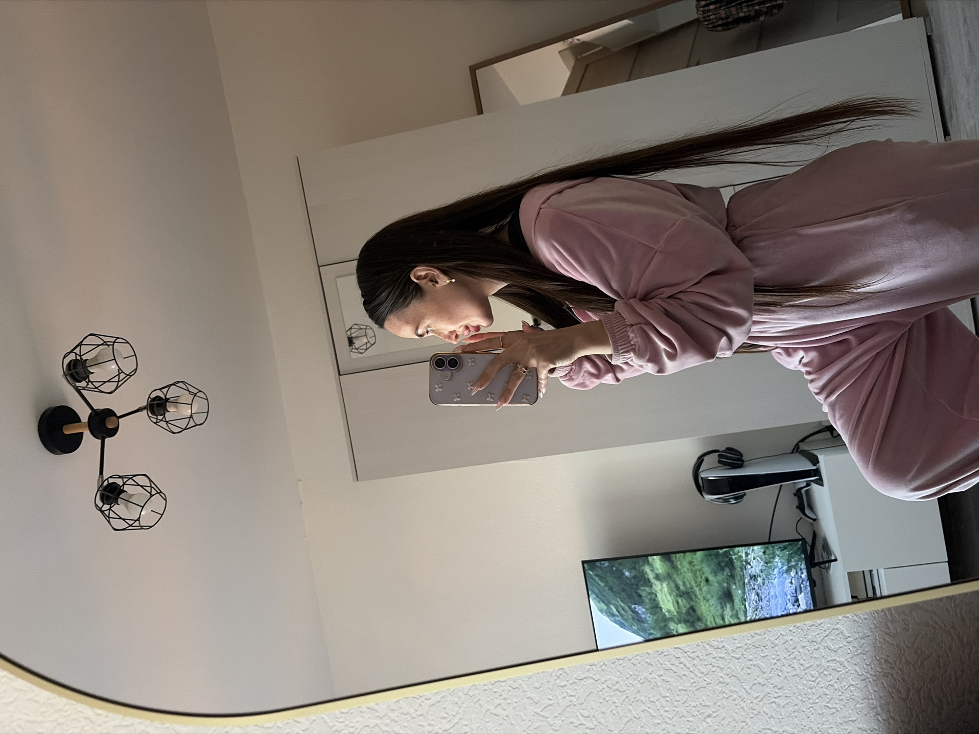 The image captures a mirror selfie taken by a person indoors. The subject, seen in profile, is a person with long, dark hair, wearing a pink long-sleeved top and matching pants, possibly a loungewear set or tracksuit. They are holding a light purple smartphone with both hands, positioned to take a picture of their reflection in a large mirror. Several rings are visible on their fingers.
The reflection in the mirror reveals parts of the room. The walls are a light, neutral color. To the left, a modern wall-mounted light fixture with three exposed bulbs enclosed in black geometric cages is visible. On the right, a television screen is reflected, displaying a nature scene with green foliage and what appears to be a waterfall or river. Below the television, a pair of white and black headphones rests on a stand. A white cabinet or piece of furniture is also reflected in the background. The mirror itself has a thin, light-colored frame. The overall setting suggests a contemporary indoor space, likely a bedroom or living area. No specific time of day or outdoor weather conditions can be determined from the image. There is no visible text.