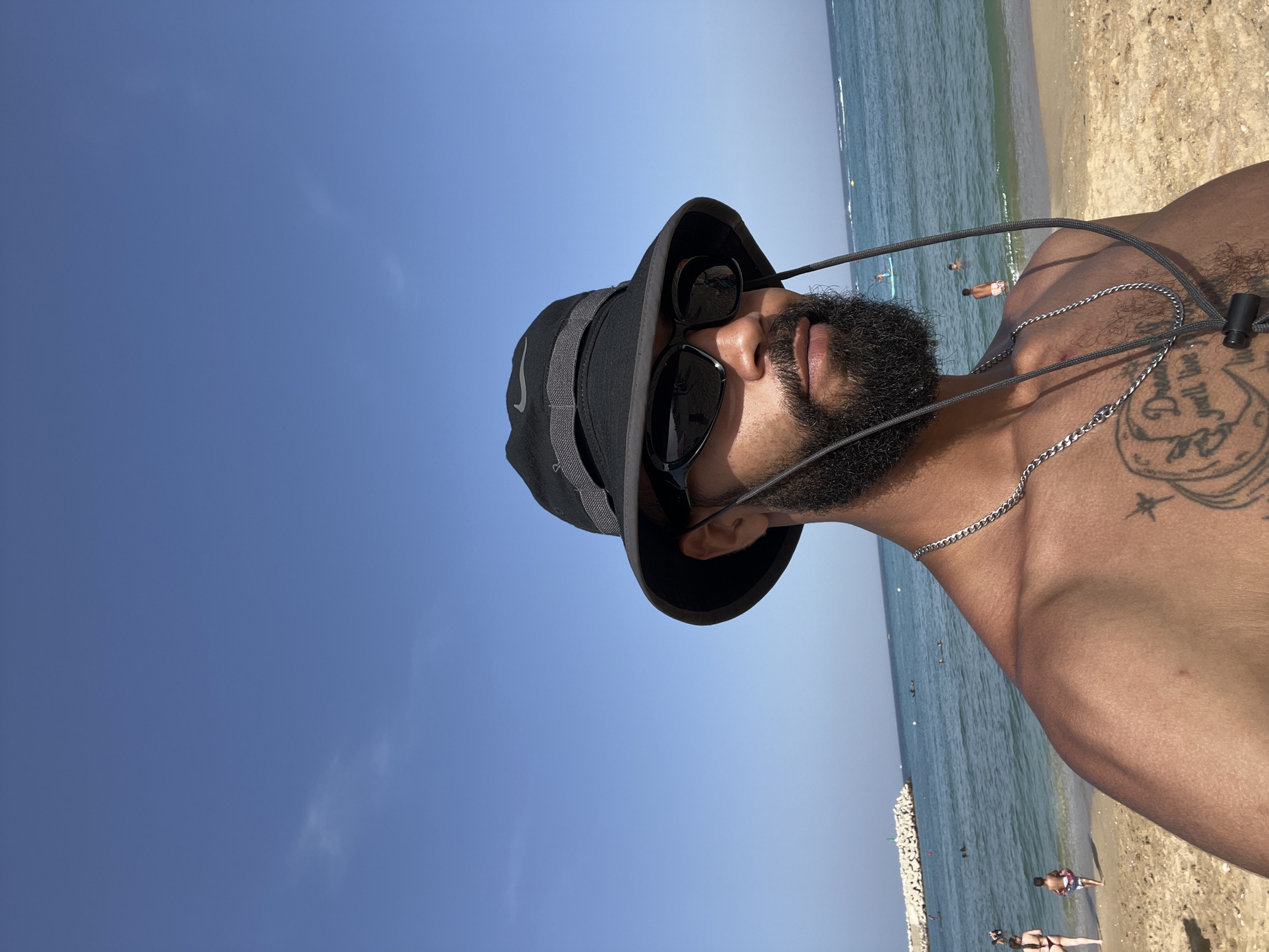 This is a selfie taken by a man on a beach, likely in M'Diq, Morocco, as indicated by the location context. The image is oriented vertically with the subject's head in the upper left portion. The man is wearing a black bucket hat and dark sunglasses, with a beard and mustache visible. He is shirtless, and a silver chain necklace is around his neck. Tattoos are visible on his chest and shoulder.
The background shows a clear blue sky above and the ocean to the right, with a sandy beach. The water is calm and blue, with several people visible in the distance, some swimming or wading. The sunlight suggests it is daytime, likely during the morning or afternoon. The overall mood appears relaxed and summery. The Nike logo is visible on the man's hat. No other significant text is clearly discernible in the image.