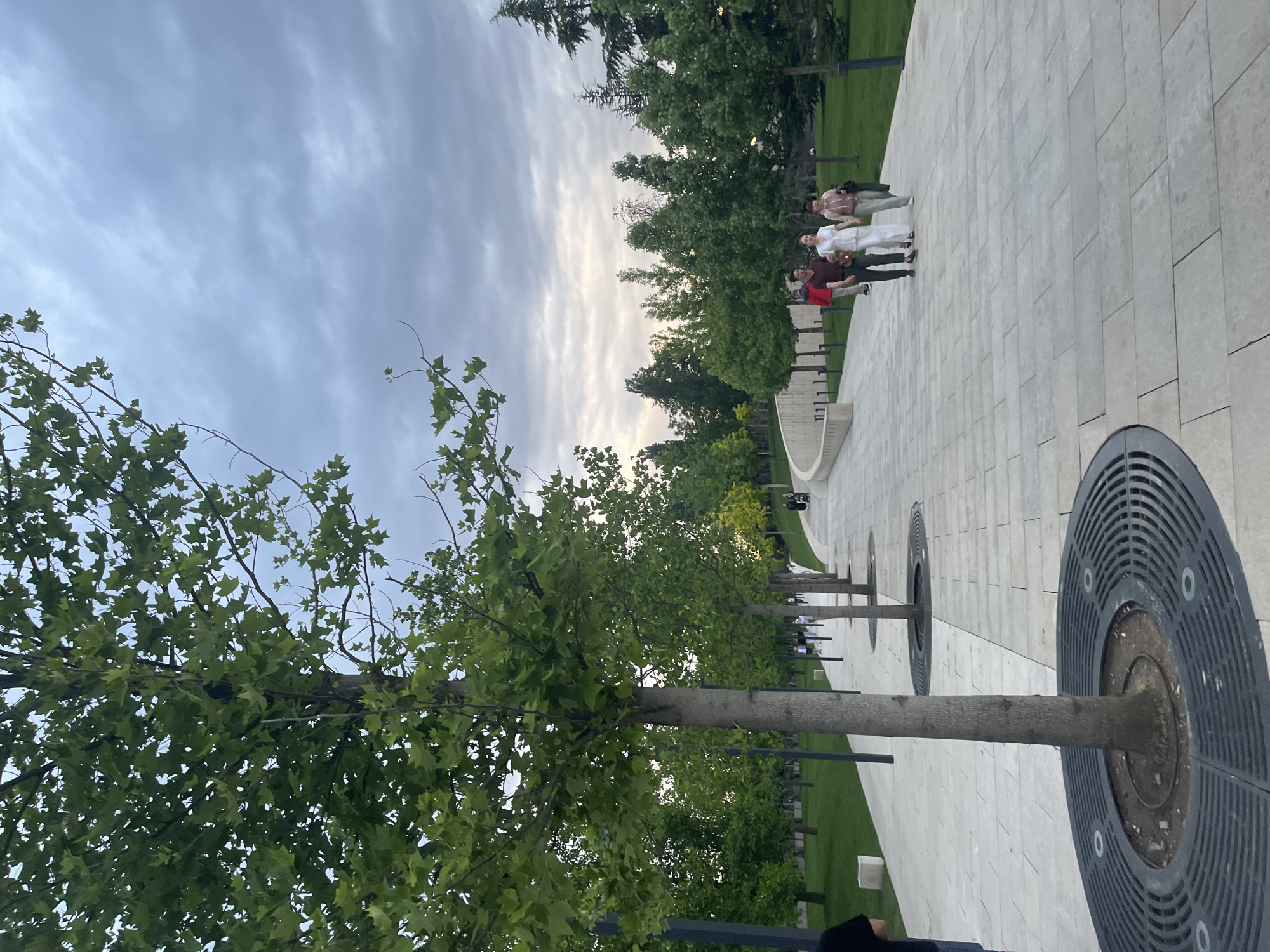 The image depicts a park scene in Krasnodar, Russia. The focus is on a paved pathway lined with young trees, each encased in a circular metal grate at its base. Four people stand on the path in the distance. The sky is overcast.