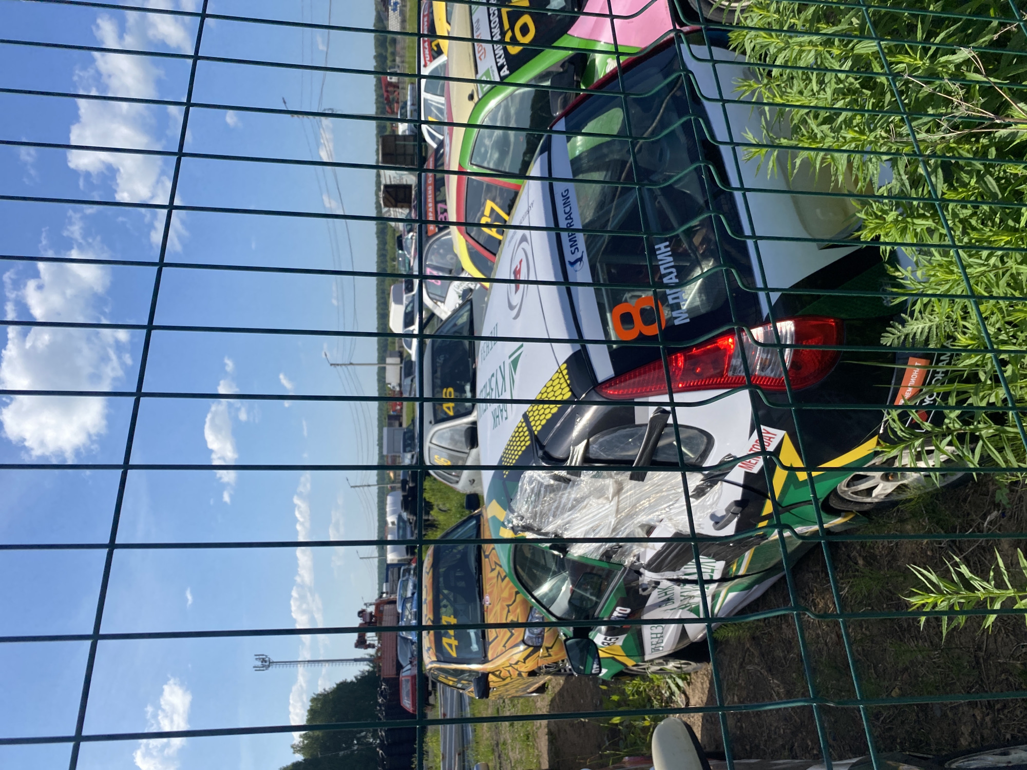 The image shows a view through a green wire fence of several race cars parked outdoors. The cars are decorated with racing livery, including numbers and sponsor logos. In the background, there are trees, a cell tower, and other vehicles, suggesting an event or a racetrack. The sky is blue with scattered clouds. The ground is dirt and overgrown with grass. The overall impression is a sunny day at an outdoor automotive event.