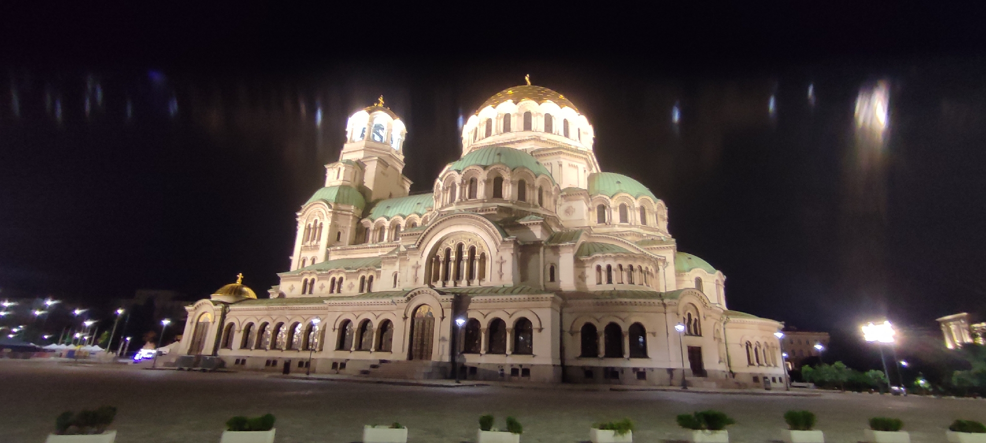 The image captures the Alexander Nevsky Cathedral in Sofia, Bulgaria, at night. The cathedral is brightly lit, highlighting its architectural features, including the golden domes and green roofs. The sky is dark, with lens flare present. In the foreground, a paved square is visible with small planters lined up. Streetlights illuminate the surrounding area. There are no people visible in the image. Other buildings are visible in the background, lit by streetlights.