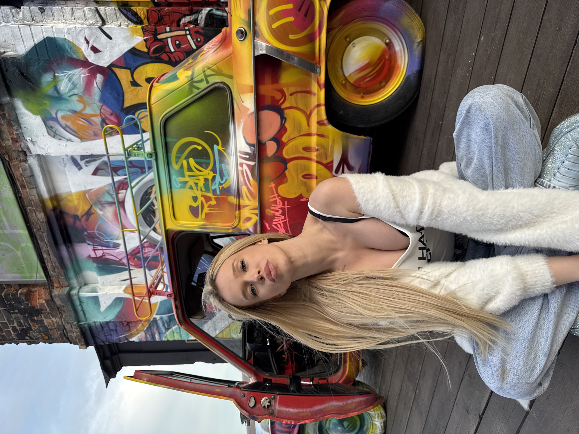 A blonde woman is sitting on a wooden deck in front of a brightly colored, graffiti-covered vintage car. The car has its driver's side door open, revealing the interior. The woman is wearing a white tank top, a fluffy white cardigan, and light-wash jeans. She has blonde hair that is partially covering her face. The background shows a brick building also covered in graffiti. The sky is overcast. The image appears to have been taken outdoors in Saint Petersburg, Russia.
