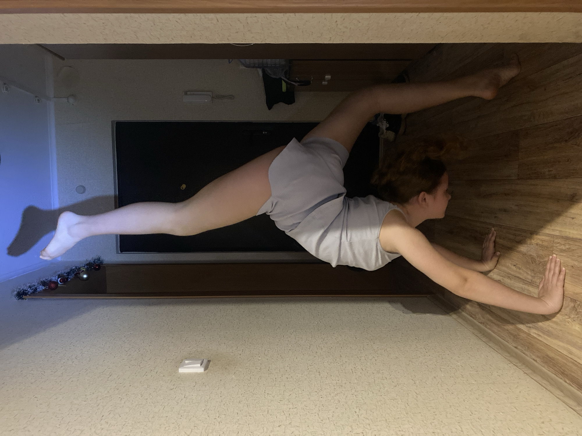 A young woman with reddish-brown hair is performing a split against a wall. Her hands are flat against the wood-paneled wall, and one leg is extended towards the left, and the other is extended towards the right. She is wearing a light-colored tank top and shorts. The setting appears to be an indoor space with patterned wallpaper, wood trim, and wood flooring. A television is visible in the background. A Christmas garland is visible on a shelf.