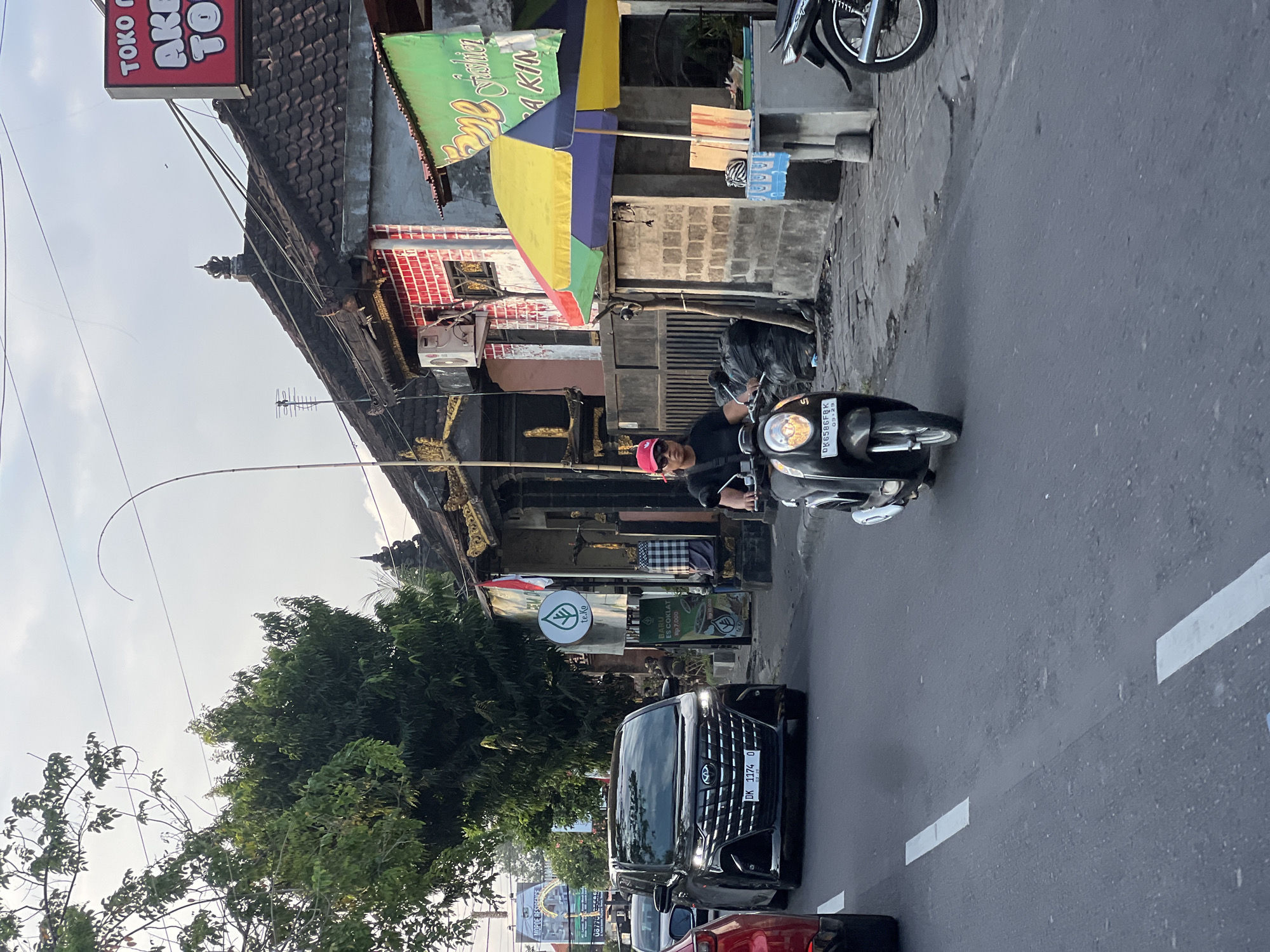 A black Toyota Avanza is driving on a road in Denpasar, Indonesia. The car has a license plate that reads "DK 1174 O". A man wearing a red hat and black t-shirt is riding a black scooter behind the Avanza. There are buildings and trees on the side of the road. One of the buildings has a red sign that says "TOKO". There is a green umbrella with yellow, purple, and blue panels. The sky is cloudy.