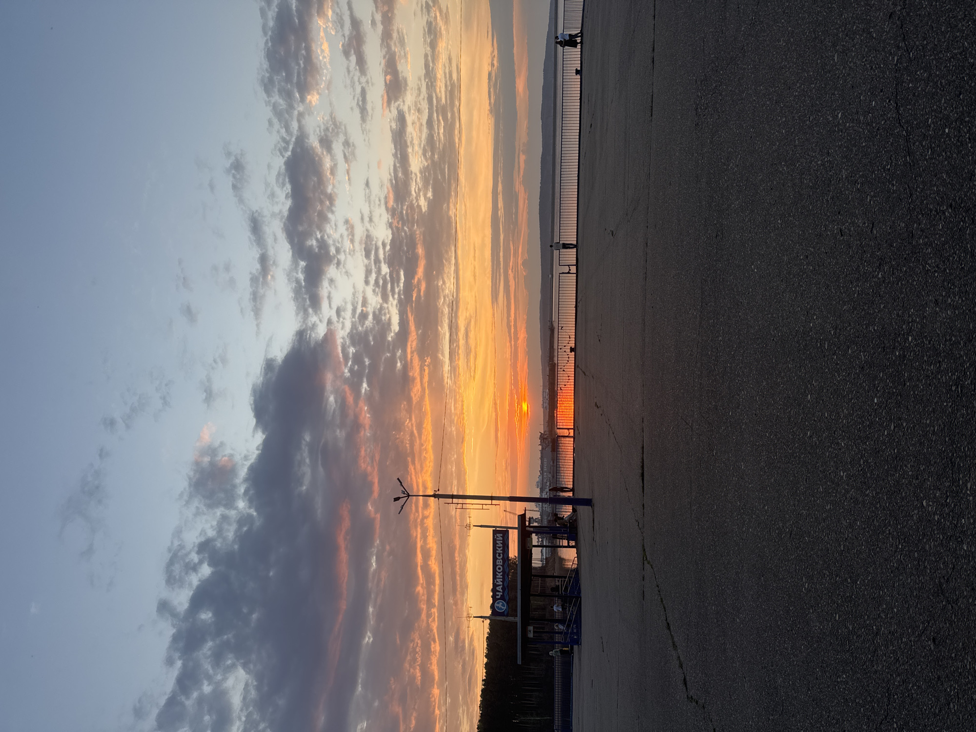 The image shows a sunset view from what appears to be a pedestrian walkway or promenade in Chaikovsky, Russia. The sky is filled with clouds, reflecting hues of orange, yellow, and pink from the setting sun. The sun itself is visible on the horizon, partially obscured by distant landmasses. A dark road or asphalt surface is the foreground, taking up a significant portion of the lower right area of the image. A metal fence runs along the edge of the walkway, separating it from the water below. Several people are silhouetted on the walkway along the edge of the water looking towards the sunset. To the left, a bus stop shelter or a sign reads "ЧАЙКОВСКИЙ" (Chaikovsky) in Cyrillic. A lamp post is close to the bus stop. Trees and some buildings are visible on the land just behind the bus stop.