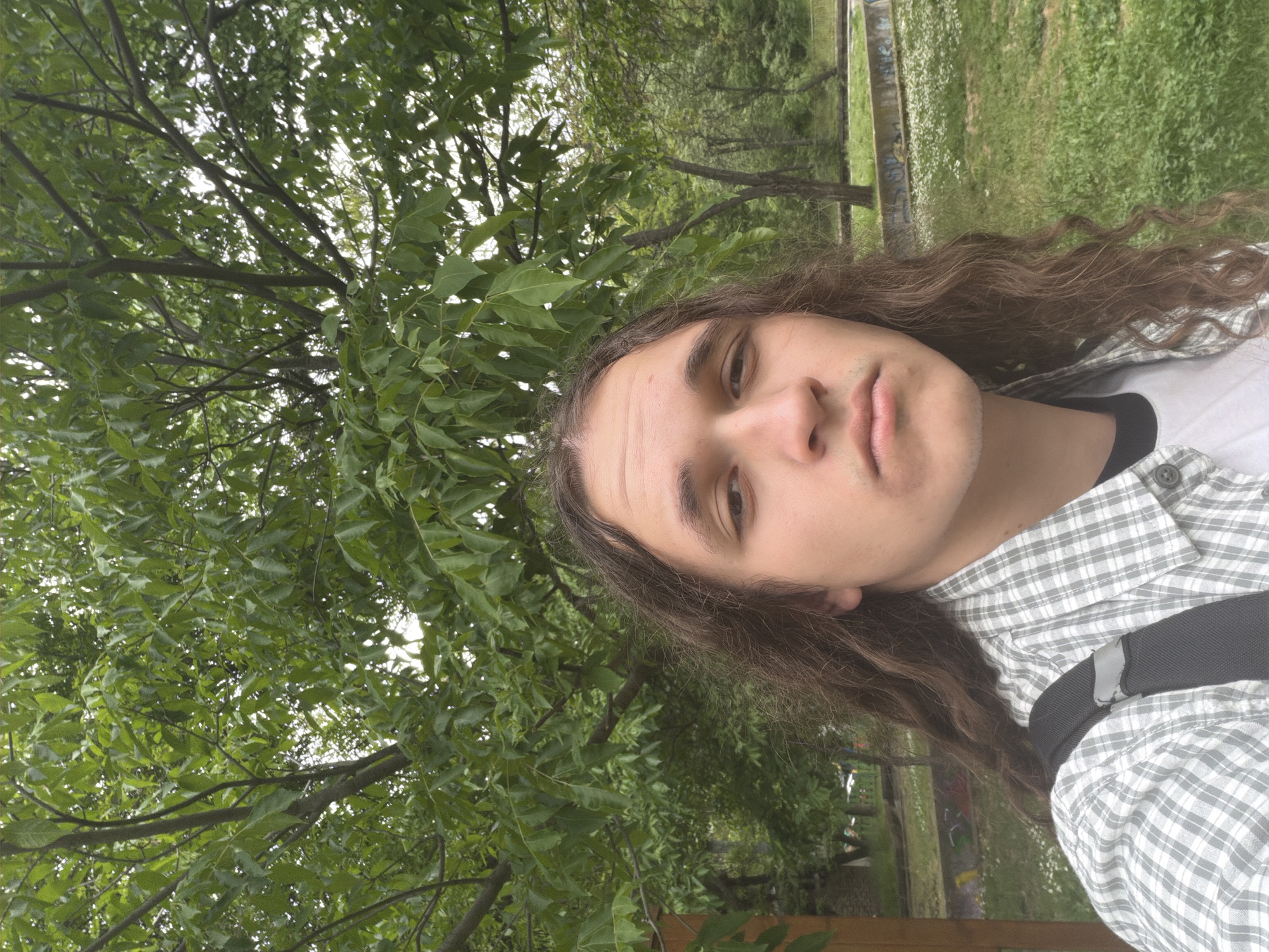 A selfie taken outdoors, likely in a park in Plovdiv, Bulgaria. A young adult with long, brown, wavy hair is the primary subject. They are wearing a light grey and white checkered shirt over a dark top, with a black strap visible across their torso. The background is filled with green foliage from trees, creating a dense, natural setting. A partially visible fence or low wall is at the bottom. In the background, there is a grassy area. The overall impression is one of a casual, outdoor scene, possibly in a park setting in the city of Plovdiv.
