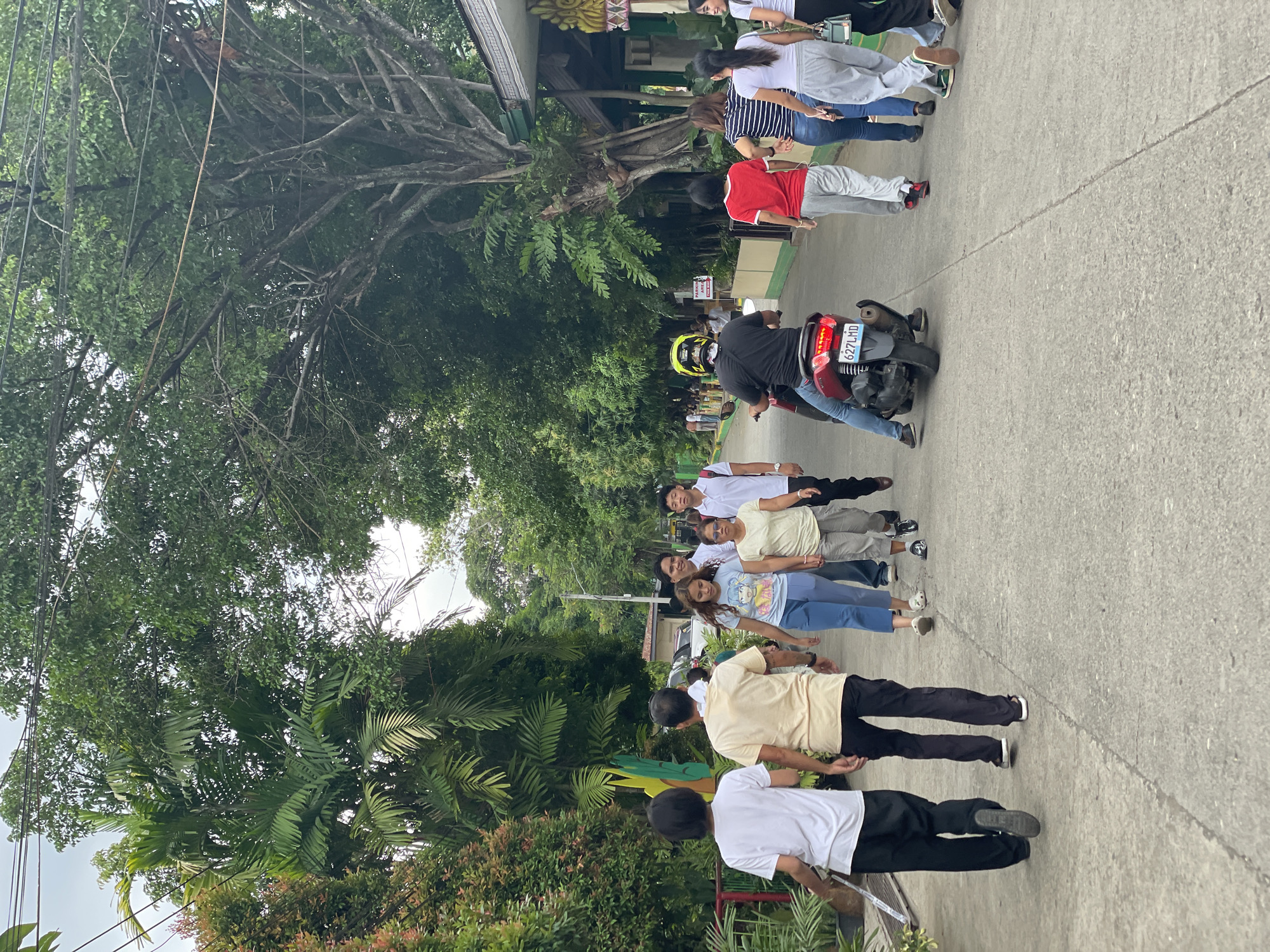 This image depicts a street scene in Davao City, Philippines, on what appears to be an overcast day. A group of people are walking along a paved road lined with lush green foliage and trees. In the foreground, on the left, two individuals are walking away from the camera, one in a white t-shirt and black pants, the other in a cream-colored t-shirt and dark pants.
Further down the road, a motorcycle carrying a rider wearing a yellow helmet is moving away from the camera. Several other individuals are walking towards the camera, including a woman in a light blue t-shirt with a graphic print and blue pants, and a woman in a white t-shirt and light gray pants. On the right side of the road, several people are walking, including a person in a red t-shirt and gray pants, and a person in a white t-shirt and gray pants.
The environment suggests a residential or mixed-use area, with buildings visible in the background, partially obscured by the dense vegetation. Power lines are visible overhead. The overall impression is a casual, daytime activity on a local street.