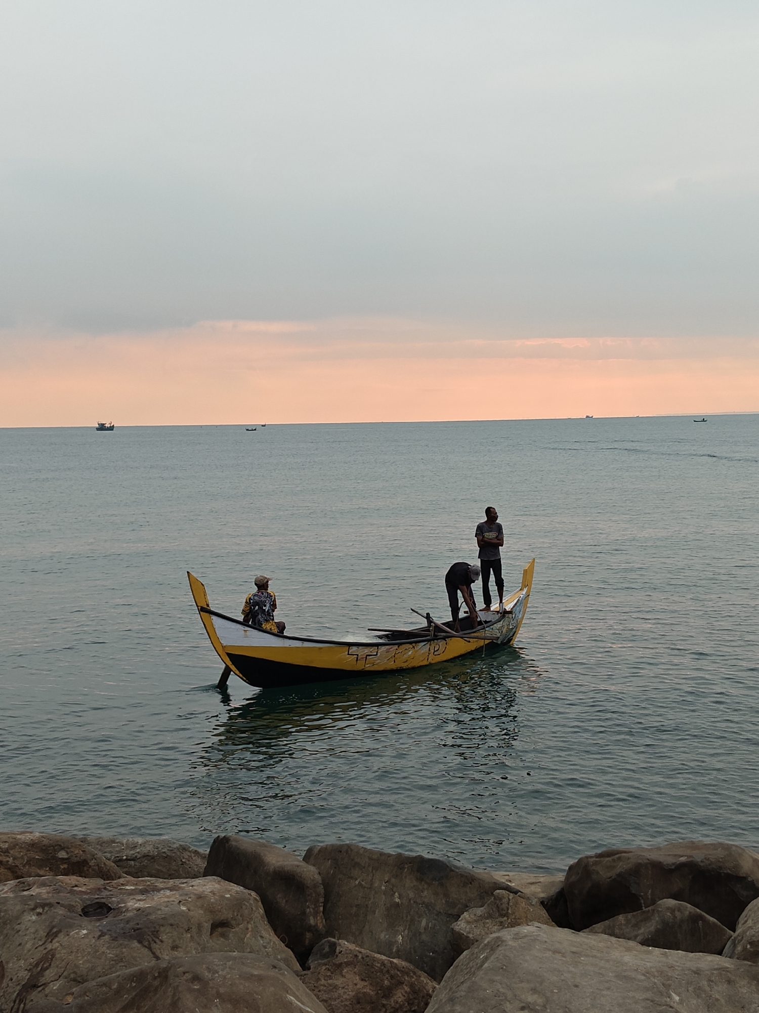 This image depicts a coastal scene in Lhokseumawe, Indonesia, at either dawn or dusk. The foreground features a cluster of large, irregular grey and brown rocks, suggesting a rocky shore or breakwater from which the photograph was taken. In the mid-ground, a traditional-style wooden boat floats on calm, dark blue-grey water. The boat has a yellow hull with a distinct black stripe along the waterline and prominently upturned, pointed bow and stern sections, also yellow. Faded white markings are visible on the hull.
Three individuals are present on the boat. Towards the bow, one person is seated, facing away from the viewer and looking towards the horizon. This individual wears a patterned shirt in yellow, black, and white, and a light-colored straw hat. Near the center of the boat, a second person stands upright, facing right (towards the stern) and observing the surroundings; they are dressed in a dark t-shirt and dark trousers. A third person is positioned slightly forward of the second, bent over, appearing to attend to something within the boat's interior, possibly equipment or contents, with oars or poles visible nearby. This individual also wears dark clothing.
The sky transitions from a light grey-blue overhead to a distinct band of orange and pink at the horizon, indicative of low sun angle. The tranquil water subtly reflects these sky colors. Several indistinct vessels are visible as dark silhouettes on the distant horizon.
