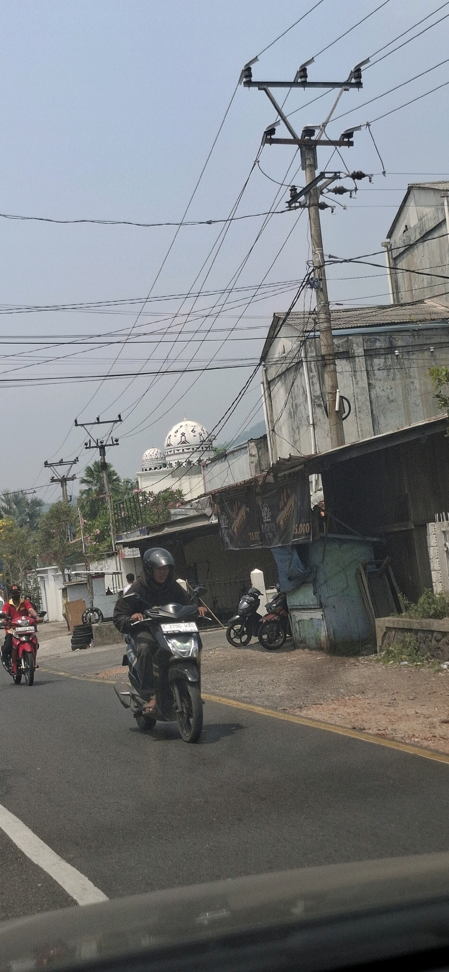 A street scene captures a motorcyclist in the foreground, wearing a dark helmet and hoodie, riding a black scooter with a license plate that reads "F 2780 UAG". To the left, another motorcyclist on a red scooter is visible. In the background, a white domed building, likely a mosque, stands out against the sky, with power lines crisscrossing overhead. A light-colored building with a corrugated roof is situated to the right of the mosque. A small, blue structure with some posters is visible in front of the buildings. The road is paved with a yellow line marking the lane. The overall environment appears to be a mix of urban and semi-rural settings, possibly in Cintaasih, Indonesia, given the presence of a mosque and the general architectural style.