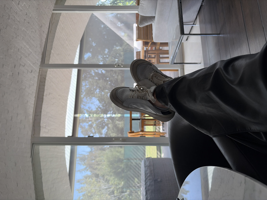 A person wearing white sneakers and dark gray pants sits in a black chair, legs extended towards a window. The window reveals a view of greenery and a blurred patio setting. A partially visible white sofa with a glass-topped table sits nearby. The scene is well-lit, with natural light streaming through the window. A brick wall is visible on the left.