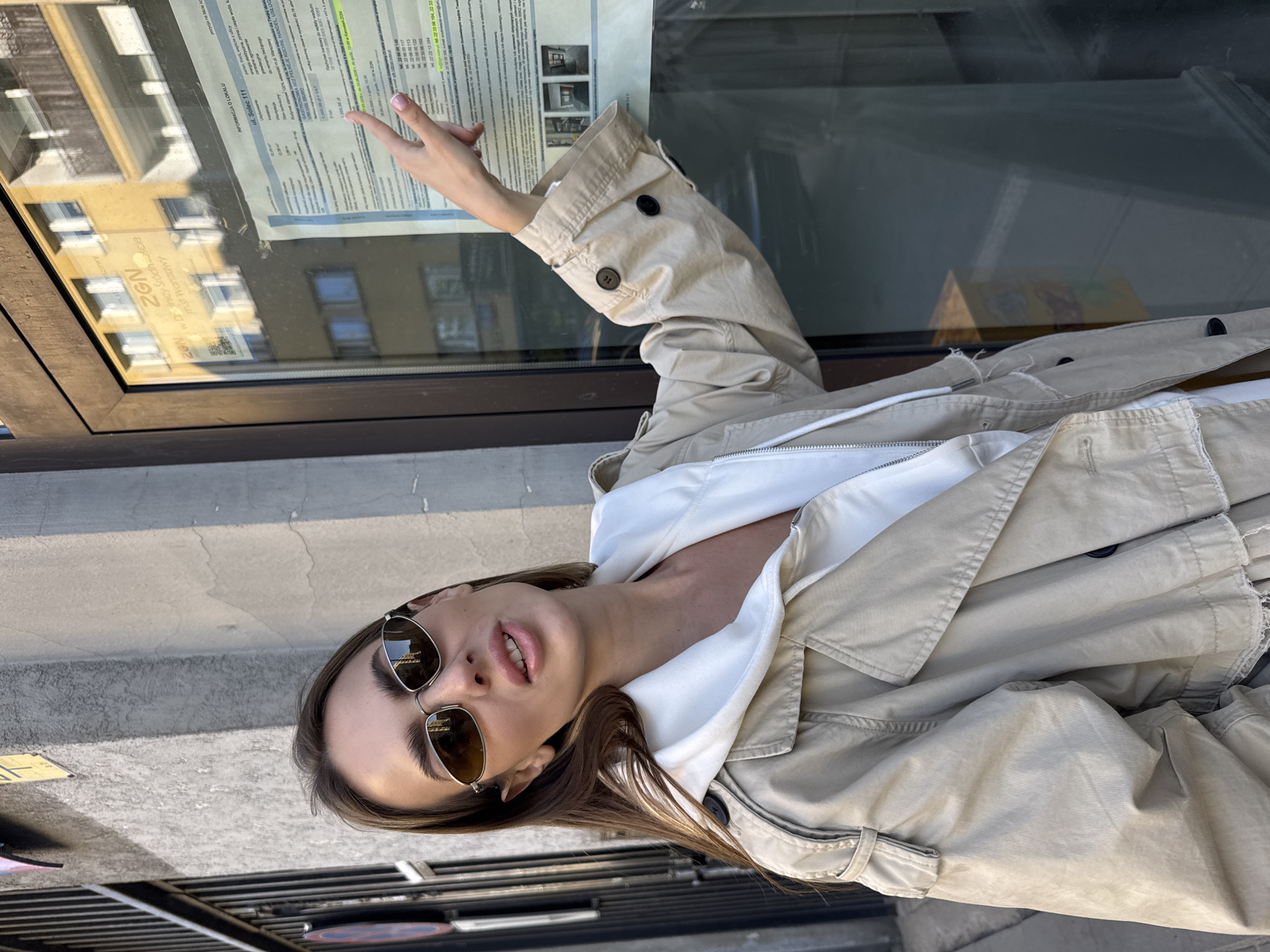 A young woman wearing sunglasses and a trench coat stands in front of a glass window in Warsaw, Poland. Her right arm is raised, pointing at a document posted on the window, likely property information for ul. Solec 111. The document is in Polish.