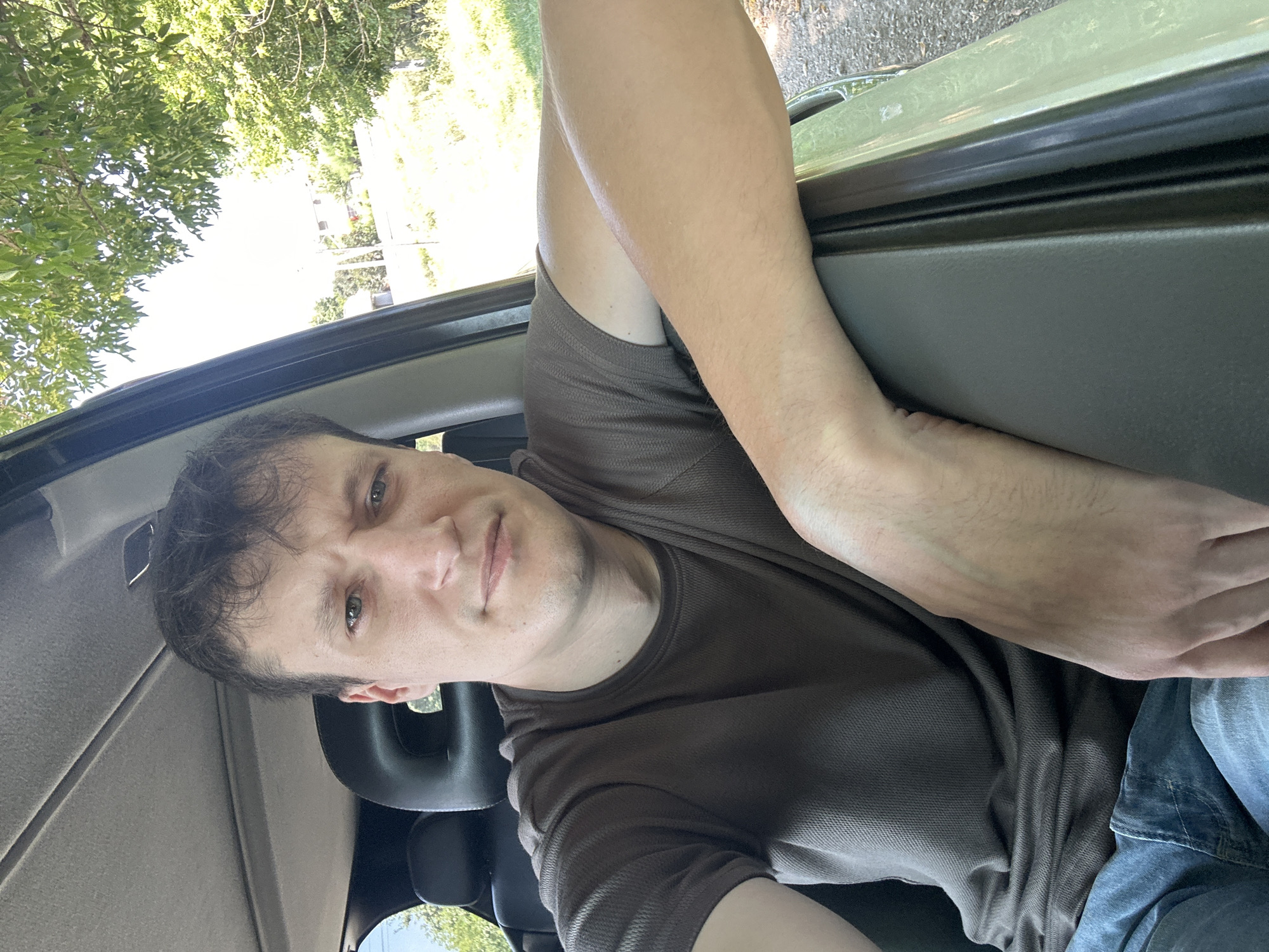 A man is sitting in the passenger seat of a vehicle, with his right arm resting on the open door frame. He is looking towards the camera, with a neutral expression. He is wearing a brown t-shirt and blue jeans. The background shows trees and a dirt road, suggesting a rural or suburban setting. The image was taken in Makiv, Ukraine.