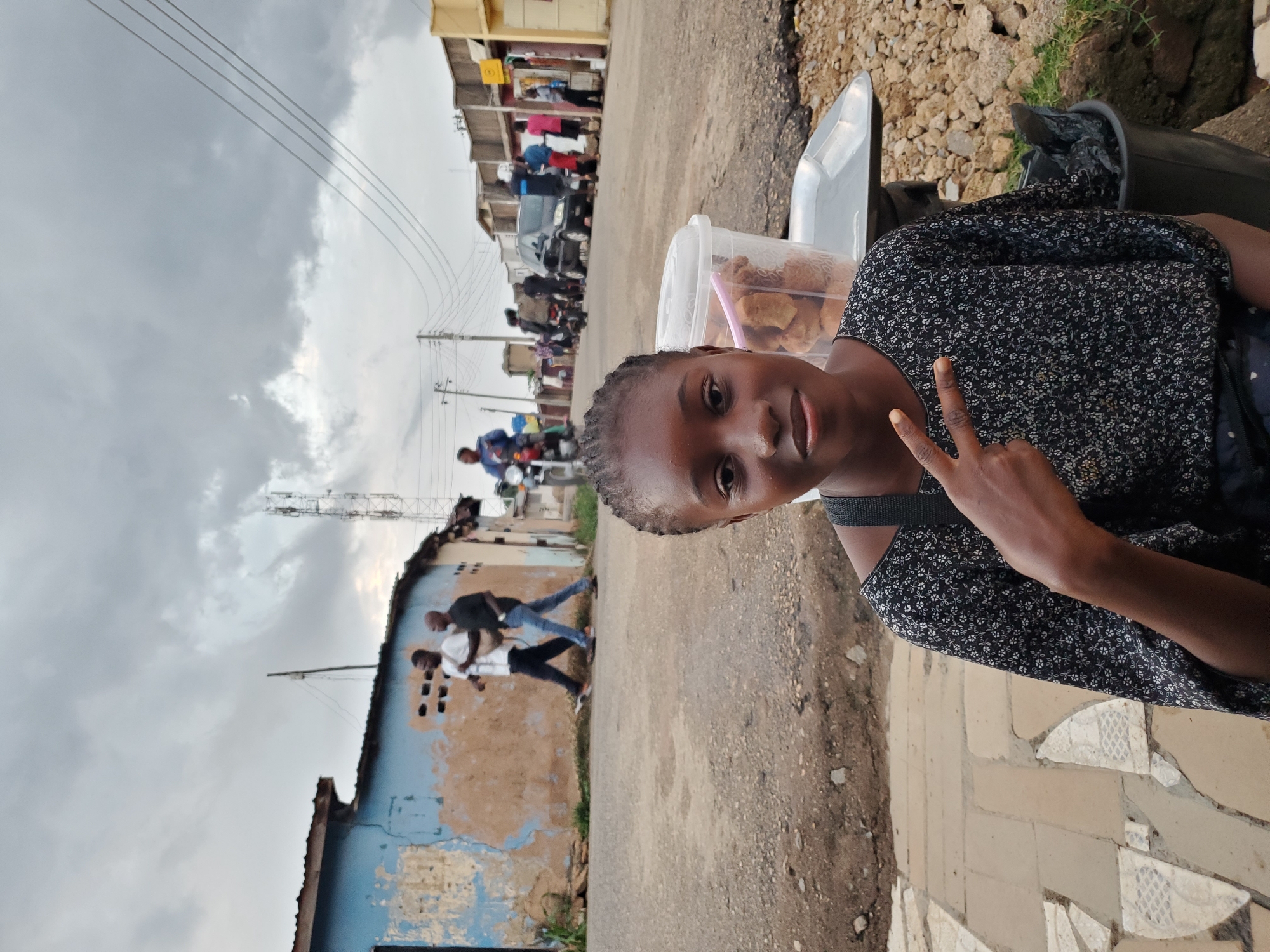The image shows a young girl making a peace sign with her right hand. She is wearing a dark-colored dress with a floral pattern. In front of her, on a white container, are some brown, fried snacks. The background depicts a street scene in Jos, Nigeria, with buildings and a cloudy sky. Several people are visible in the background, some walking and others on motorcycles. The weather appears overcast, suggesting it might be daytime with a possibility of rain. The buildings in the background show signs of wear and tear. There is no visible text in the image. The girl's expression is pleasant, and she appears to be enjoying herself.