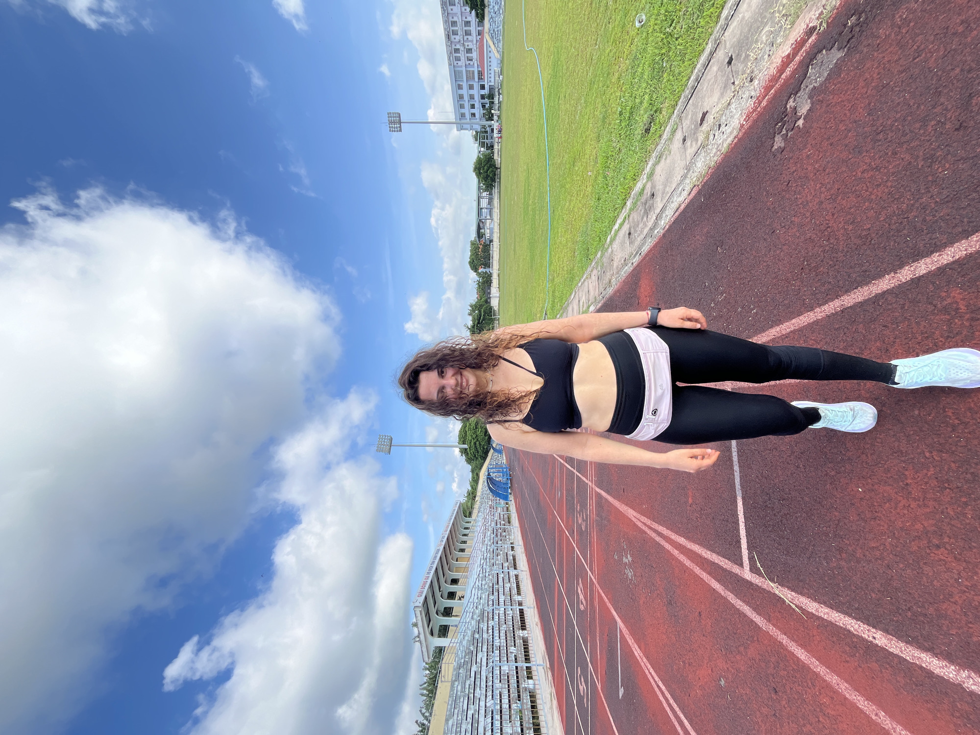 A smiling woman with curly brown hair stands on a red running track at a stadium in Phan Thiết, Vietnam. She is wearing a black sports bra, black leggings, a light pink fanny pack, and light blue athletic shoes. The running track has white lane markers. In the background, there are stadium bleachers with white seats and a green field. The sky is blue with fluffy white clouds. Lights for the stadium are visible in the distance.