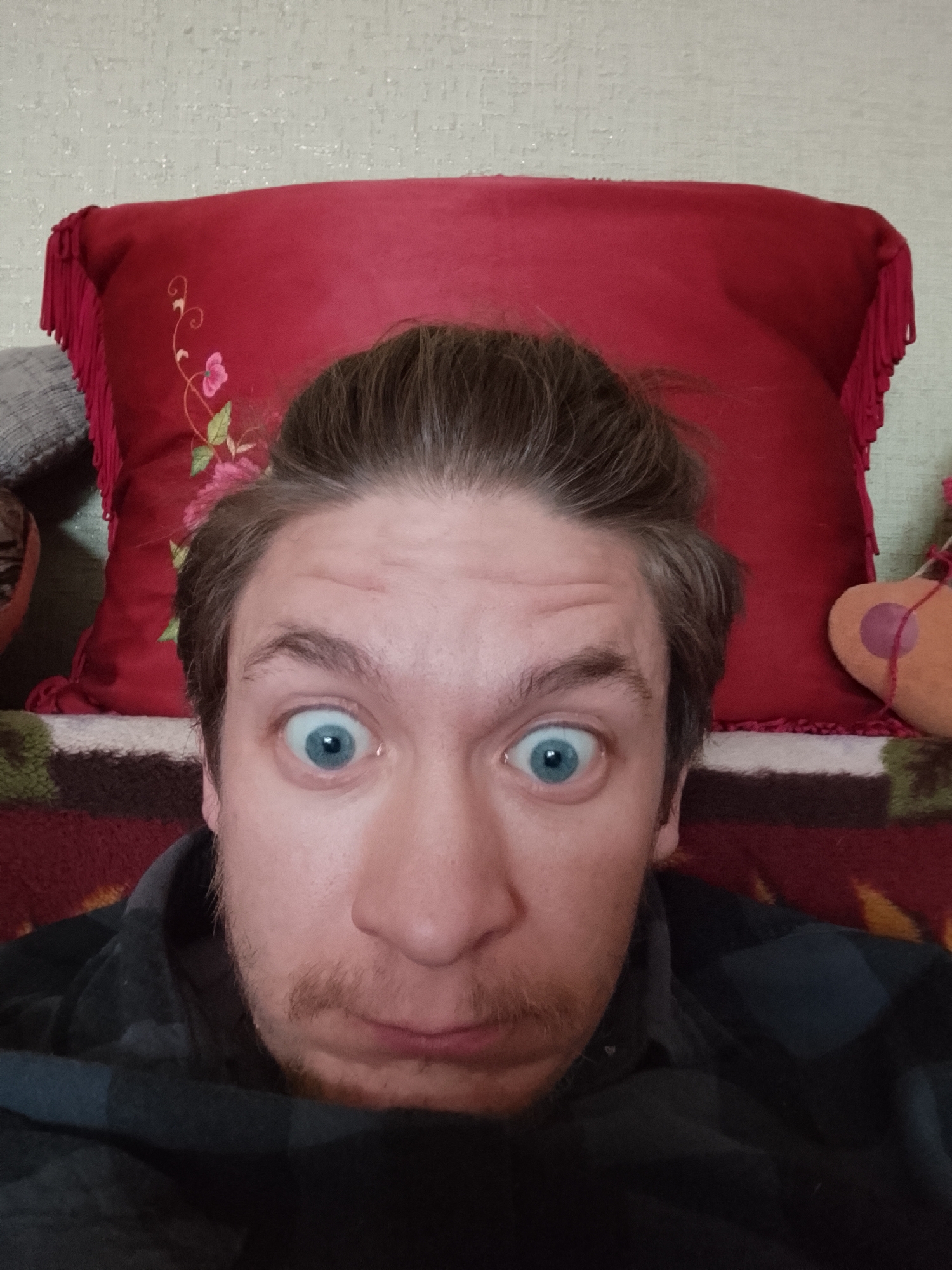 A close-up selfie depicts a man with light brown hair pulled back into a small bun, large blue eyes widened in surprise, and a small mustache. He is wearing a dark plaid shirt. Behind him is a red pillow with floral embroidery and tassels along the edge. A multicolored blanket or fabric is partially visible below the pillow. The background is a textured, light-colored wall. The location is Unknown, Unknown.