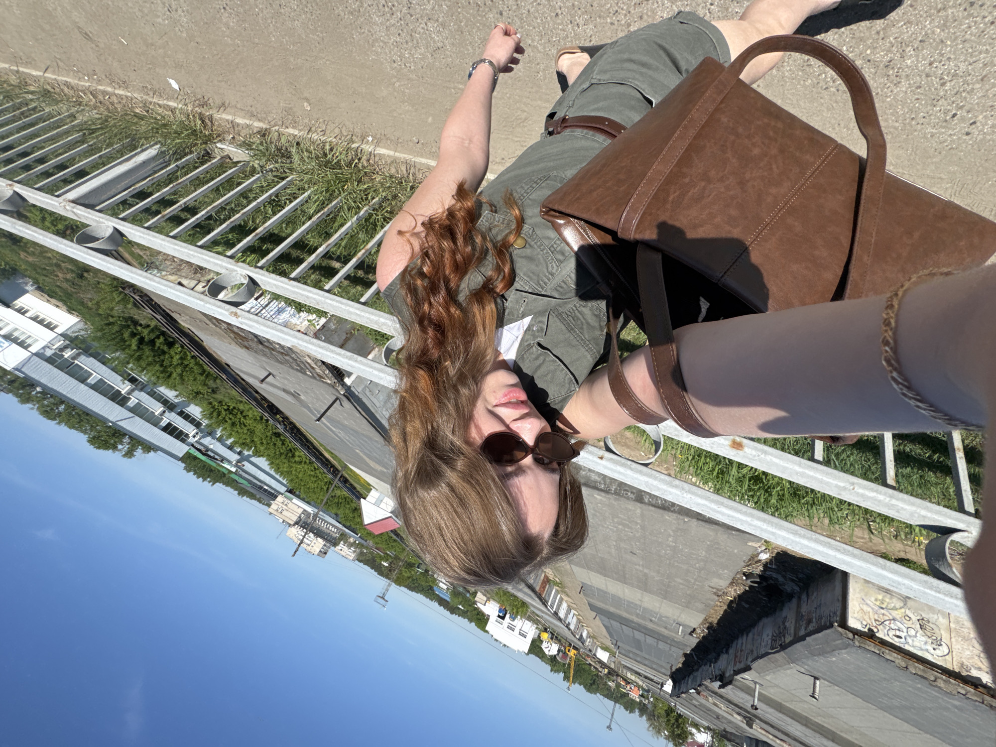 A person, seen from a high angle, lies on a concrete surface adjacent to a metal railing. The person wears sunglasses, a green outfit, and carries a brown bag. The background shows buildings, vegetation, and a clear blue sky, consistent with an urban environment, specifically the city of Chaikovsky, Russia.