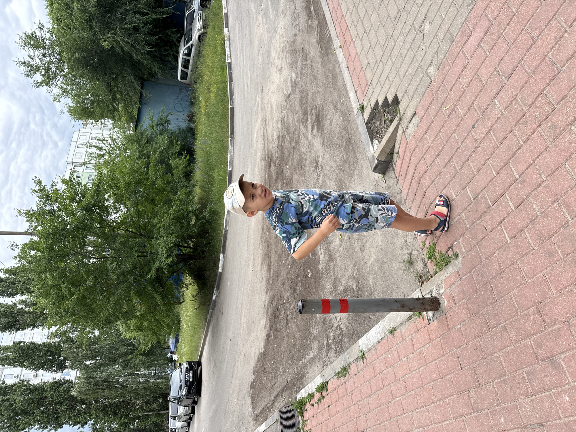 A young boy stands on a brick sidewalk in a city. He is wearing a light-colored baseball cap, a blue and green camouflage t-shirt, and matching shorts. He has a concerned expression on his face. The sidewalk is bordered by a dirt and asphalt road with parked cars and tall buildings in the background. The sky is overcast.