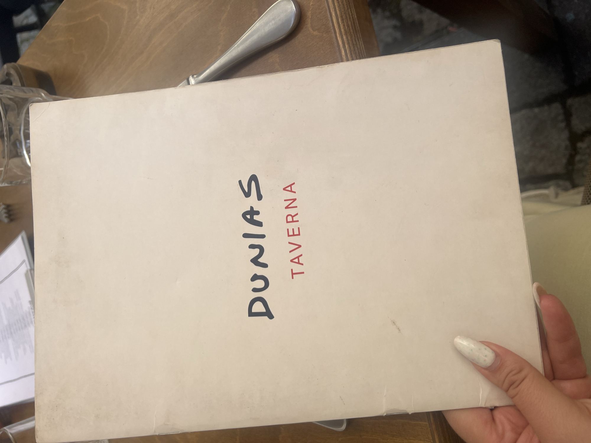 A close-up shot captures a person's hand holding a cream-colored menu at a dining establishment, identified as "Dunia's Taverna" in Plovdiv, Bulgaria. The menu features the name "DUNIAS" in large, dark, hand-drawn style capital letters, stacked vertically, with "TAVERNA" written below it in smaller, red, sans-serif capital letters, also oriented vertically.
The hand has long, white fingernails adorned with small, colorful speckles. The menu, which appears to be a booklet, shows some signs of wear and minor smudges on its surface. It rests on a light-colored wooden table, where a metal utensil (possibly a knife or fork) and a clear glass are partially visible. In the background to the left, another menu or stack of papers can be seen. To the right, hints of what resembles stone or cobblestone paving suggest an outdoor or rustic setting. The image depicts the moment of browsing a menu, likely preceding an order at the tavern.