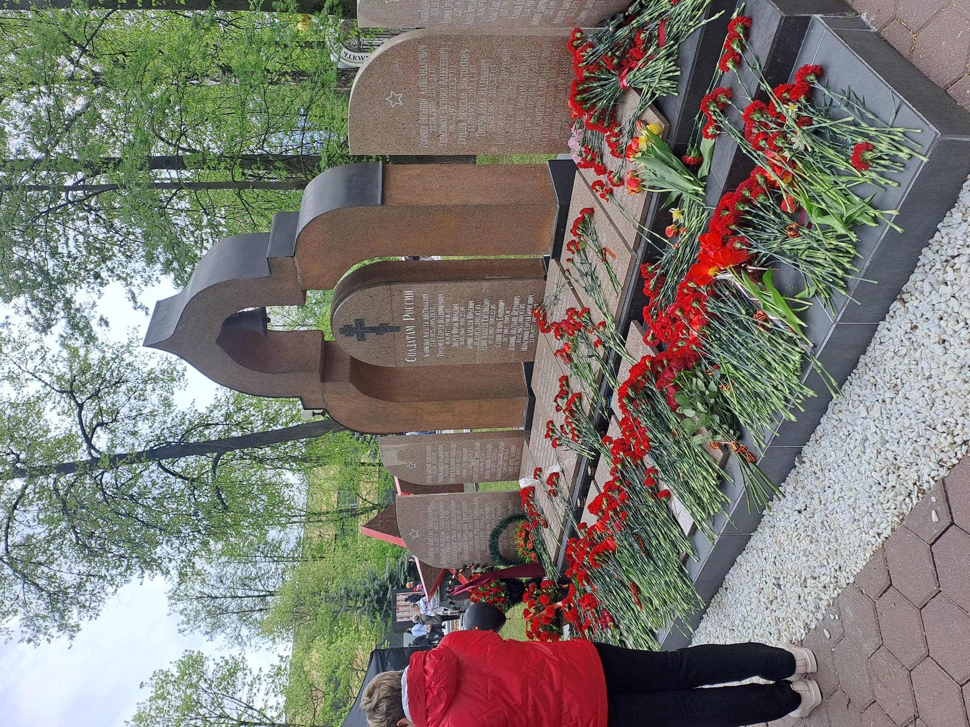 The image shows a memorial complex in Elektrostal, Russia. The memorial consists of several vertical stone plaques with engraved names and dates, likely commemorating soldiers. Many red carnations are laid at the base of the memorial. A woman in a red jacket and dark pants stands in front of the memorial. The area surrounding the memorial is paved with stone tiles and white gravel. Trees are visible in the background. Other people are seen in the distance, behind the woman in red.