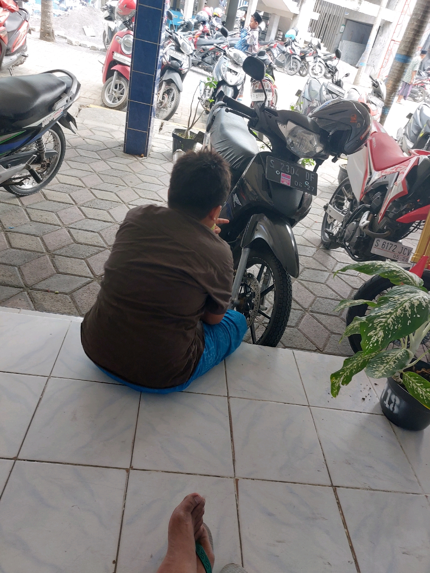 The image captures an outdoor scene in Sumberrejo, Indonesia, likely a street or parking area, filled with numerous parked motorcycles. In the foreground, a person is seated on the ground with their back to the camera, wearing a brown shirt and blue pants. They are positioned next to a black motorcycle with a license plate displaying "S 2304 DF" and a date "05-27". A black helmet rests on the handlebars of this motorcycle. To the right, another motorcycle with a red and white color scheme is partially visible, its license plate reading "S 6172 JCV" and "05-28". A potted plant with broad, variegated leaves is situated to the right of the seated person. The ground is tiled with a geometric pattern, and in the background, more motorcycles and a building structure are visible. The overall atmosphere suggests a typical street scene in a populated area of Indonesia.