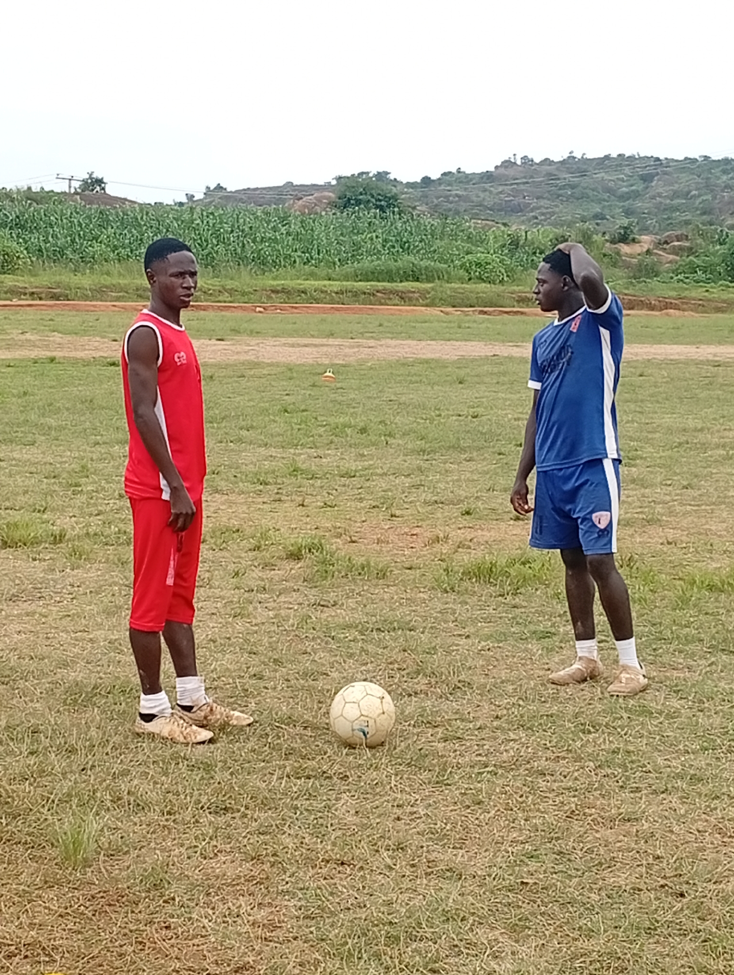 Two young men, likely in their late teens or early twenties, stand on a grassy field with a soccer ball between them. One man, on the left, wears a red sleeveless jersey and red shorts. The other man, on the right, wears a blue t-shirt with white trim and blue shorts. Both are wearing athletic shoes. They appear to be in a rural or semi-rural area, with a line of green crops in the background, followed by hills with sparse vegetation and rocky outcrops. The sky is overcast. This scene could be from a sports training ground or a recreational area in or around the city of Jos, Nigeria, known for its cooler climate and hilly terrain.