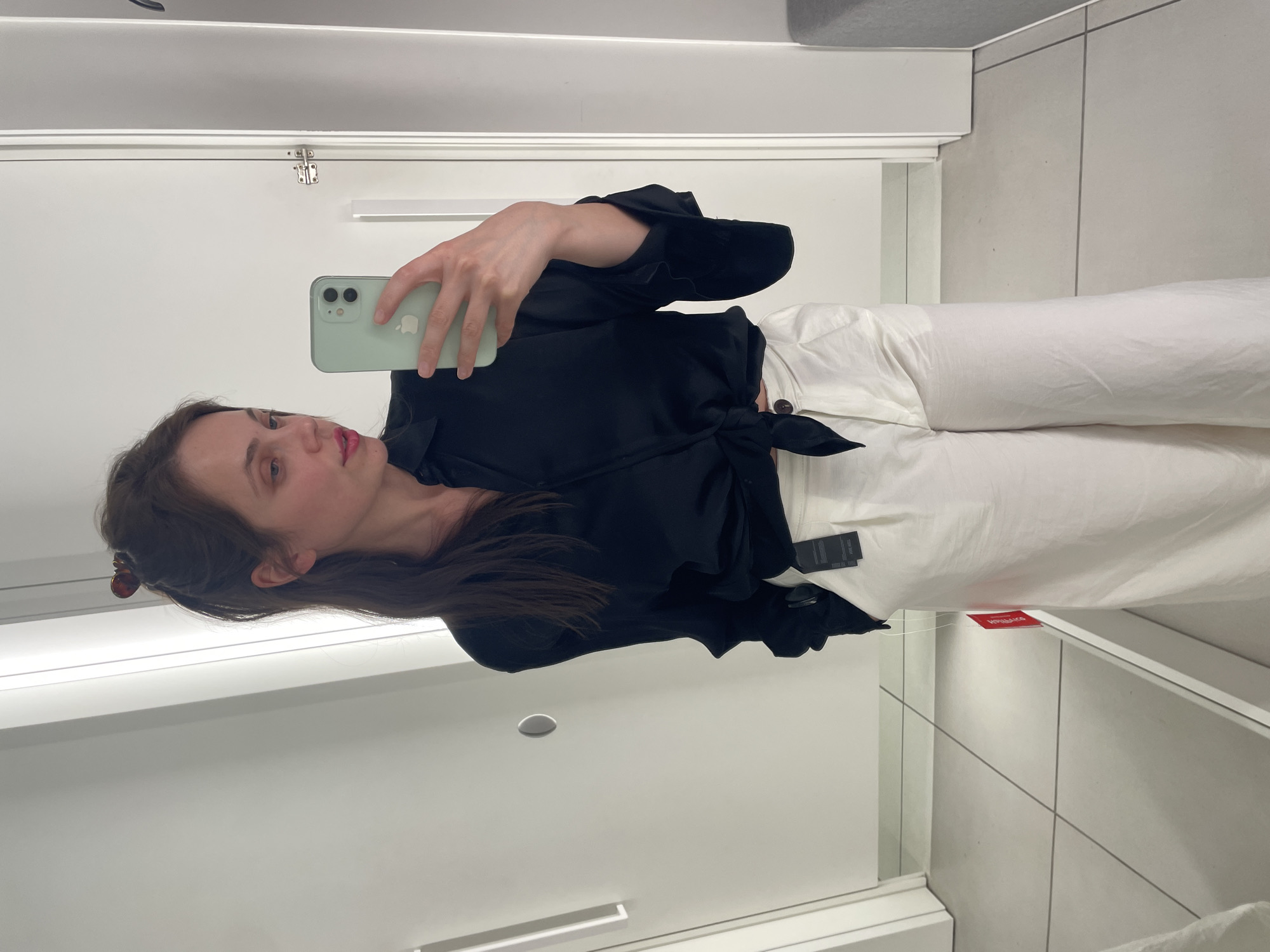 A woman with dark hair pulled back in a messy bun, wearing a black knotted blouse and white wide-leg pants, stands in a brightly lit, tiled fitting room. She is taking a mirror selfie with a light green smartphone. Tags are visible on her pants. The room features white walls and doors, with a gray bench visible to the right. The image was captured in a fitting room located in the city of Vecsés, Hungary.