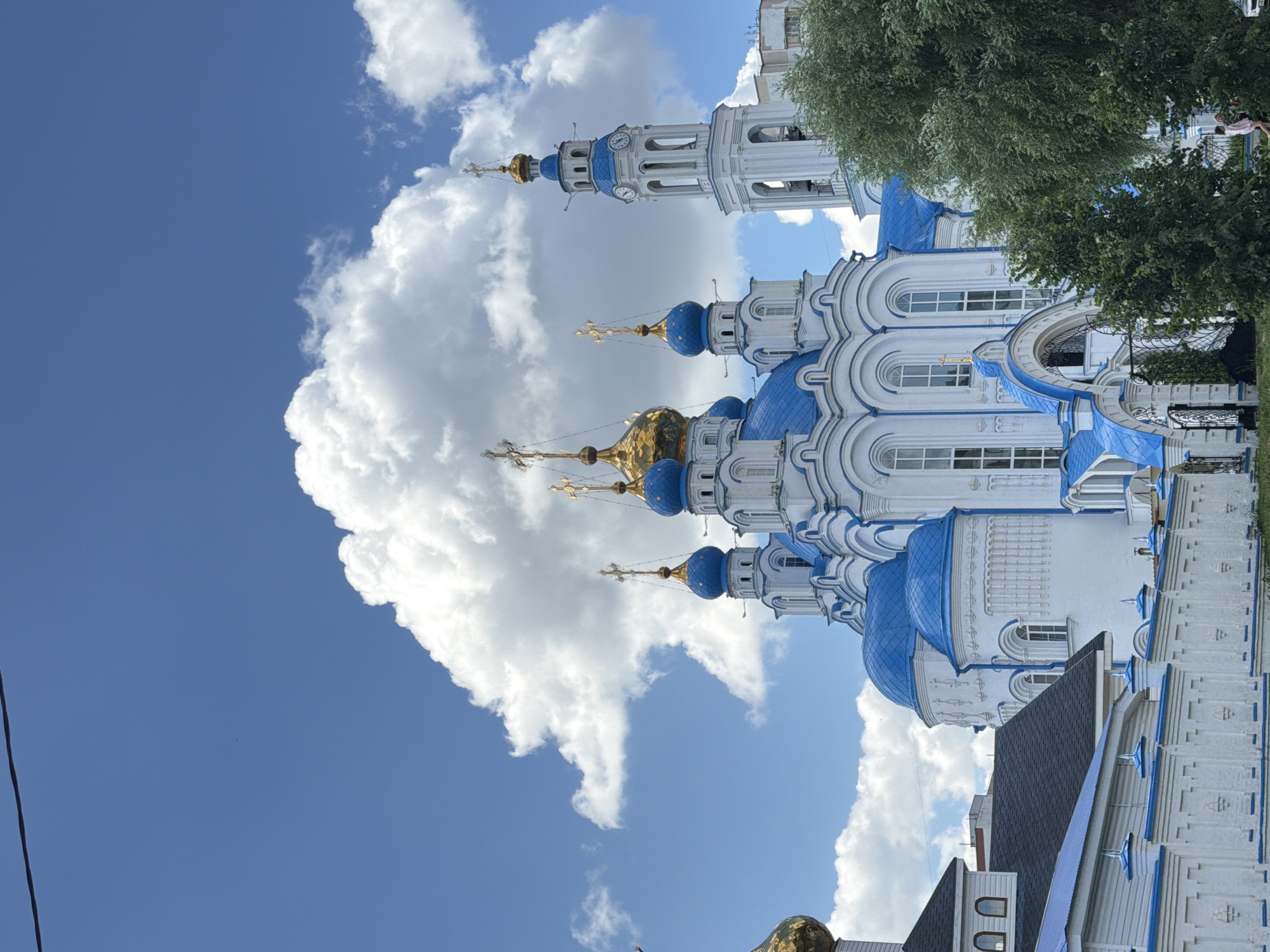 A low-angle medium shot captures a white and blue domed cathedral against a bright blue sky dotted with fluffy white clouds. The cathedral features multiple blue domes topped with golden crosses, and a tall white bell tower with a blue cap and clock faces. The building is adorned with white architectural details. In the foreground, a white picket fence with blue accents separates the viewer from the cathedral grounds. A lush green tree partially obscures the right side of the cathedral. Two individuals, one in a pink shirt and shorts and another in a darker outfit, are visible walking near the fence in the lower right corner of the frame. The overall scene depicts a serene and picturesque view of a religious landmark.