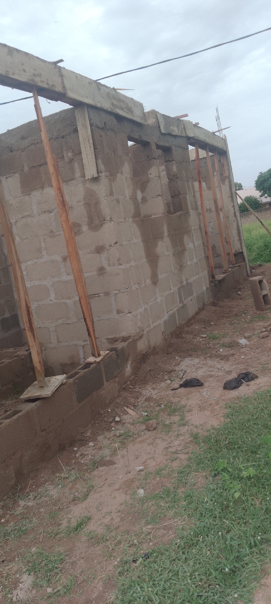 A construction site in Jalingo, Nigeria, shows the partially built walls of a structure made of cinder blocks. Wooden beams are propped up by vertical wooden posts, serving as temporary support for the ceiling or roof structure. Some of the walls appear to be wet, suggesting recent concrete work or rain. The ground is a mixture of dirt and sparse grass. In the foreground, two dark, lumpy objects, possibly plastic bags filled with debris or waste, lie on the ground. A single cinder block sits upright on the ground to the right of the structure. Overhead, a wire runs across the sky, which is overcast. In the background, trees and the roofs of other buildings are visible.