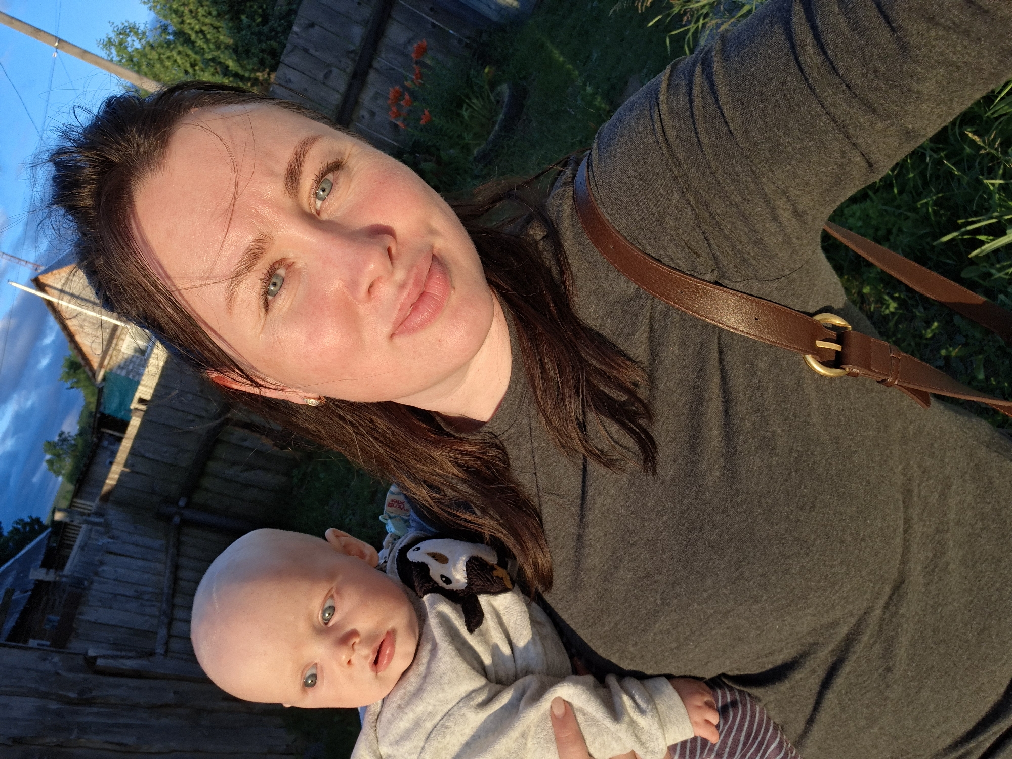 This is a medium close-up selfie of a woman holding a baby. The woman, with brown hair and blue eyes, is wearing a gray t-shirt and a brown leather strap is visible over her shoulder. The baby, with light skin and blue eyes, is wearing a gray sweatshirt with a penguin applique and striped pants. They are standing outdoors in a rural setting. In the background, there is a wooden fence, some buildings, trees, and green grass. The sky is visible with a hint of blue and clouds. The lighting suggests it is either early morning or late afternoon. The location is identified as the city of Karsha, Russia.