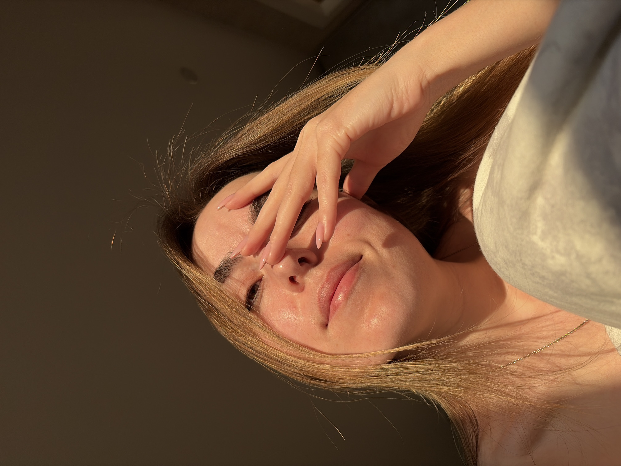 The image depicts a woman with long, light brown hair partially obscuring her face. Her hand is raised, covering part of her forehead and right eye. She has fair skin, pink nails, and is wearing a light-colored garment and a delicate gold necklace. The background is a dark, indistinct space with some areas illuminated by a warm light source, creating strong shadows. The photograph is taken from a low angle, looking upward at the subject. The location cannot be determined from the image alone, and it is not possible to determine if the image was taken in Izhevsk, Russia.