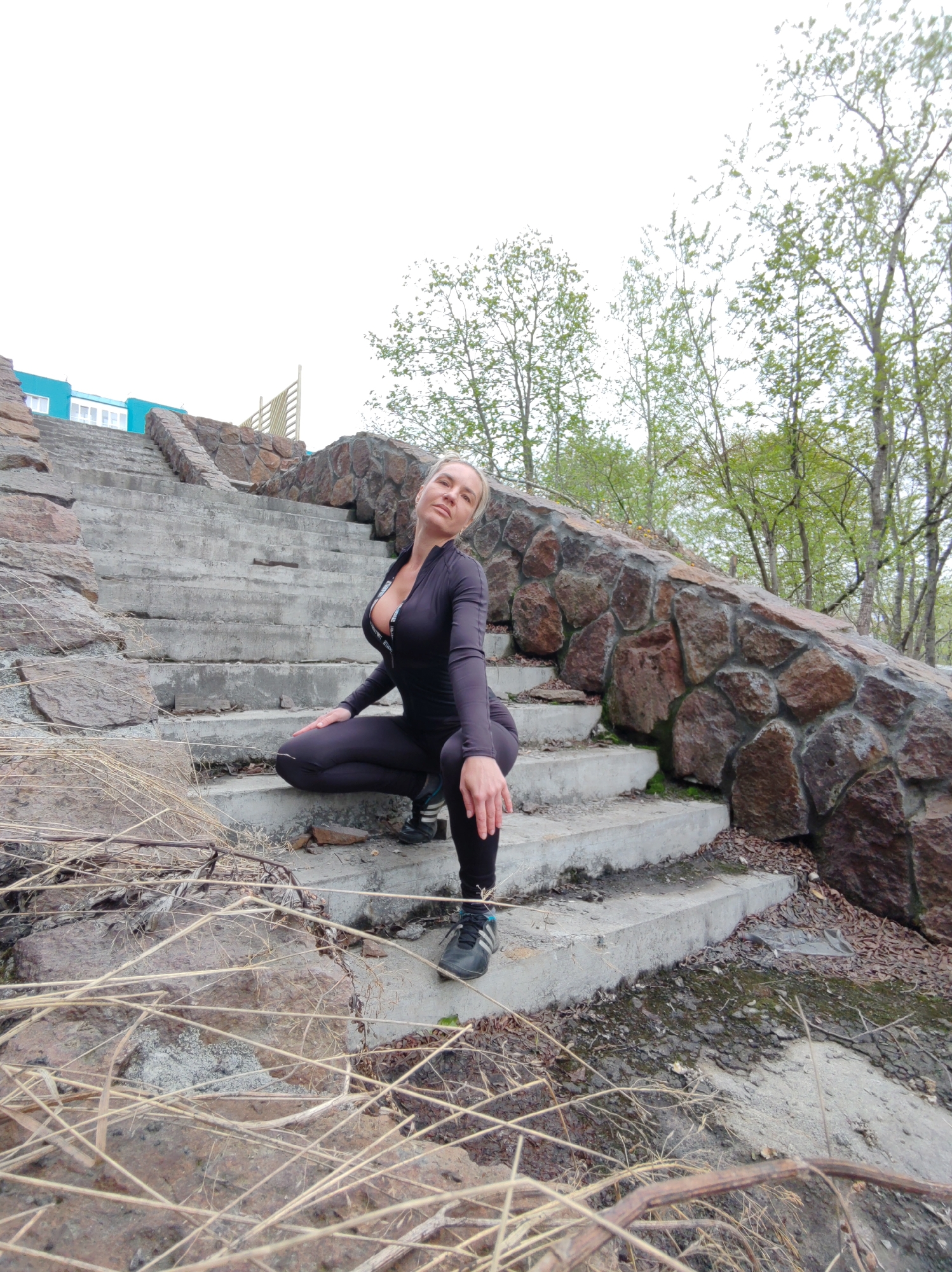 A blonde woman wearing a dark, form-fitting athletic outfit is crouching on a concrete staircase with a stone retaining wall to her right. She is looking towards the camera with her left hand resting on her knee and her right arm bent at the elbow. The staircase appears to be made of weathered concrete and is bordered by rough-hewn stones. Dry, straw-like vegetation is visible in the foreground and on the steps. In the background, several trees with sparse green foliage are visible against a bright, overcast sky. A blue building with white windows is partially visible at the top of the stairs to the left. The scene appears to be an outdoor urban setting.