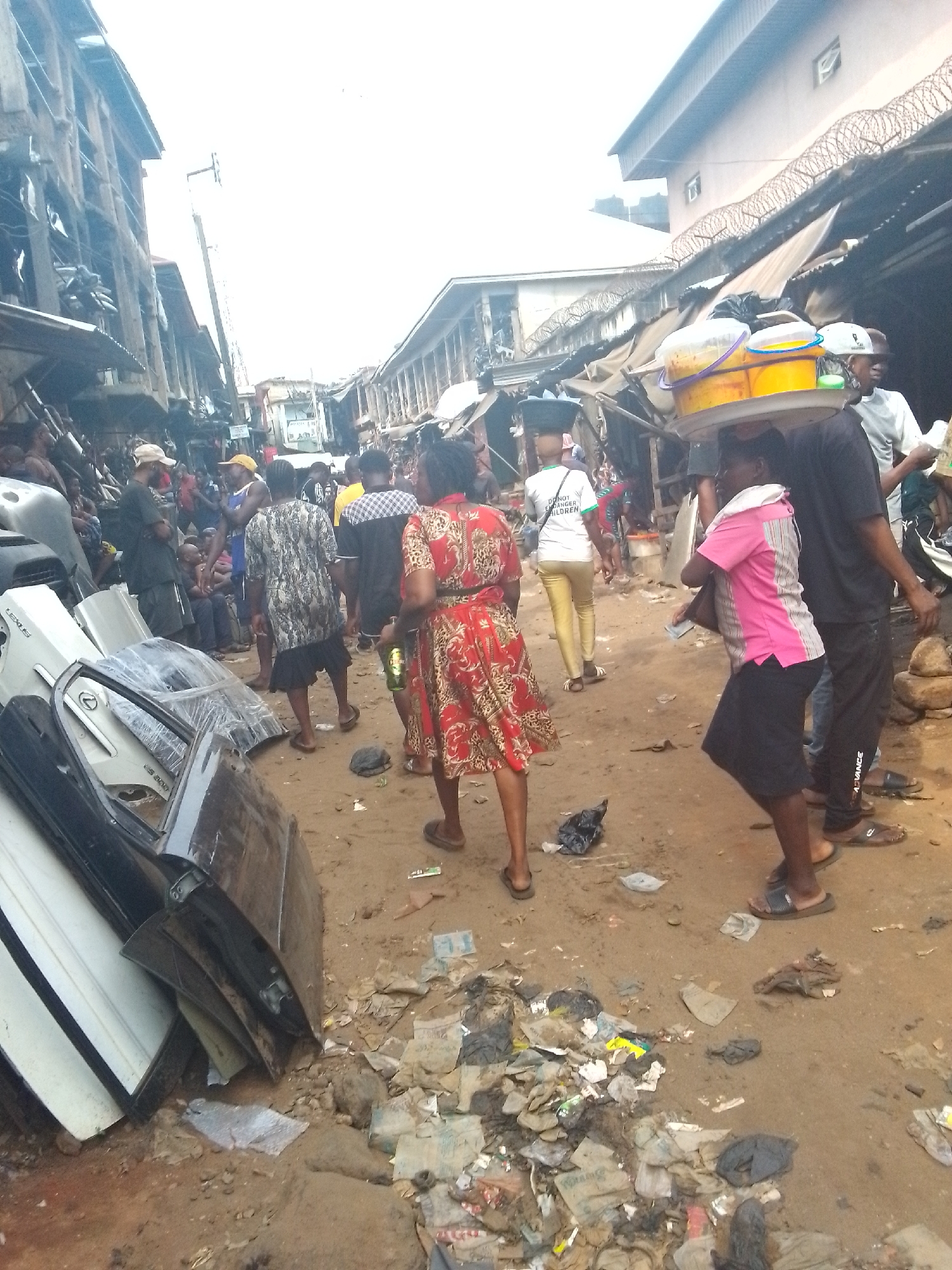 This image depicts a bustling outdoor market scene, likely in Onitsha, Nigeria, given the context. The unpaved ground is a mix of dirt and scattered refuse, including plastic bags, paper, and other debris.
Numerous people are visible, moving through the narrow street. Many are dressed in casual clothes. In the foreground on the left, a pile of assorted car parts, including doors and bumpers, is stacked. A white car door, possibly a Lexus, shows the partial inscription "LEXUS LS 500".
Towards the center-right, a vendor carries a large, flat tray on their head, laden with a yellow bucket, a white bucket, and other containers, suggesting food items or goods for sale. Next to them, a person in a pink and white striped shirt walks with a handbag. Further back, another person's white t-shirt bears partial text "DONOR...CHILDREN", and black pants of another individual have "ADVANCE" printed on them.
The street is lined with market stalls or shops, many with corrugated metal roofs and rough structures. Some buildings in the background are taller, and parts of their perimeter walls feature barbed wire. The scene is brightly lit, indicating daytime, though direct sunlight isn't casting sharp shadows, suggesting it might be an overcast day. The overall atmosphere is one of lively commerce and daily activity in a crowded, urban setting.