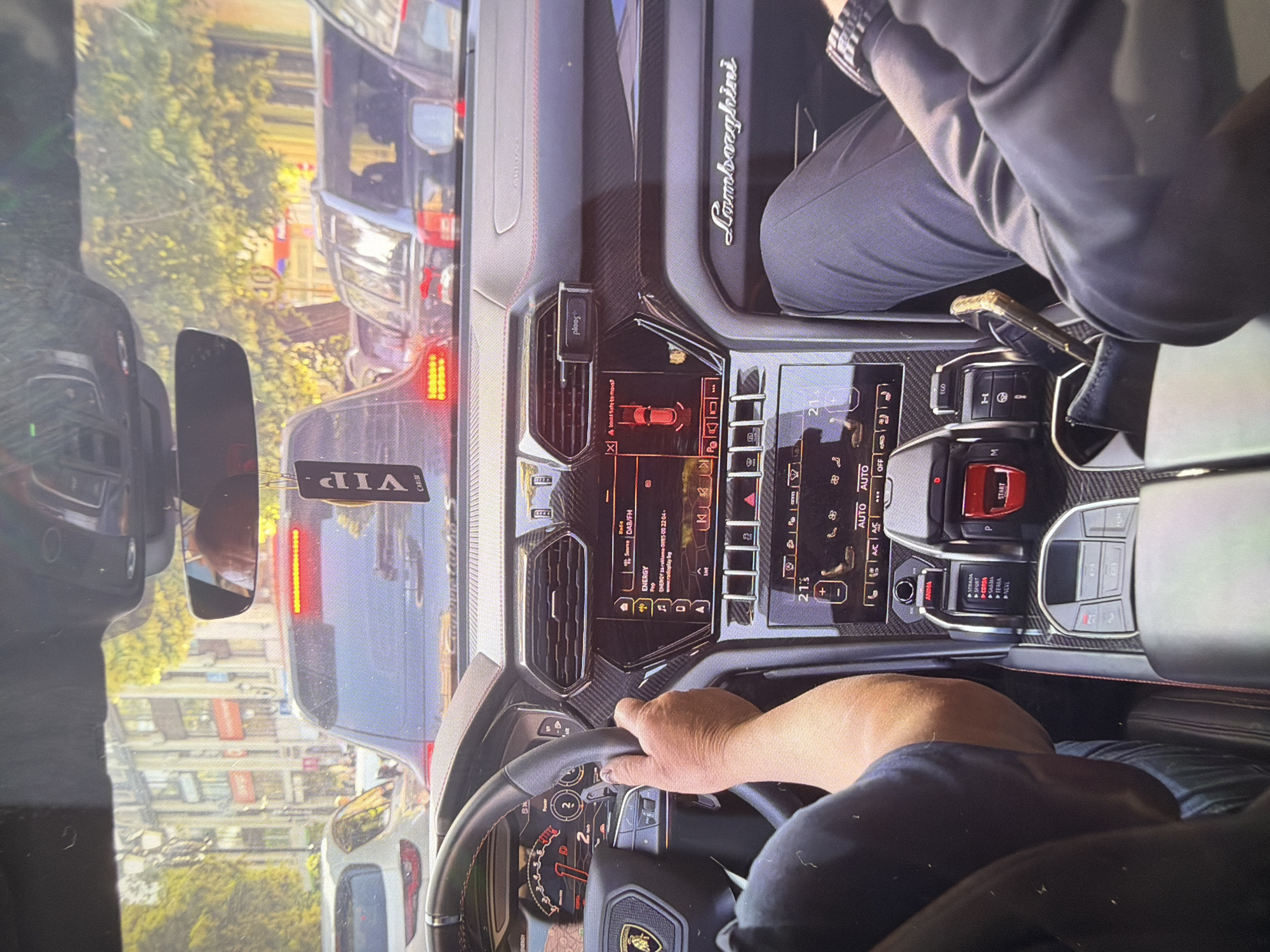 The image captures the interior of a luxurious black Lamborghini Urus, seen from a slightly elevated perspective within the vehicle, looking towards the front. A driver, identifiable by their arm and hand on the steering wheel and a blue shirt, is operating the car. A passenger's legs and arm, dressed in a gray long-sleeved top and dark trousers, are visible on the right.
The car is in traffic during what appears to be a bright, sunny day, indicated by the light filtering through the windshield. Outside the car, other vehicles are visible, including a dark SUV directly in front with illuminated red brake lights and a "VIP CAR2021" tag hanging from its rear-view mirror. Beyond the vehicles, trees and buildings suggest an urban or suburban setting.
The car's highly detailed dashboard features extensive carbon fiber trim. Multiple digital screens display information; the main infotainment screen shows "ENERGY" playing on "DAB/FM" from "radioplay.bg", with a time of "21:5" and a temperature of "21.5" degrees Celsius. Another screen shows a car outline and a warning. Below the screens, the distinctive Lamborghini Urus controls are visible, including a red "START" button and the ANIMA (Adaptive Network Intelligent Management) drive mode selector, with options like "STRADA", "SPORT", "CORSA", "SABBIA", "TERRA", and "NEVE" (road, sport, race, sand, off-road, snow). A phone is docked in a "pleases" branded mount on the air vent. The "Lamborghini" script is prominently displayed on the passenger side of the dashboard.
The location context, Dermantsi, Bulgaria, aligns with the "radioplay.bg" visible on the infotainment screen.