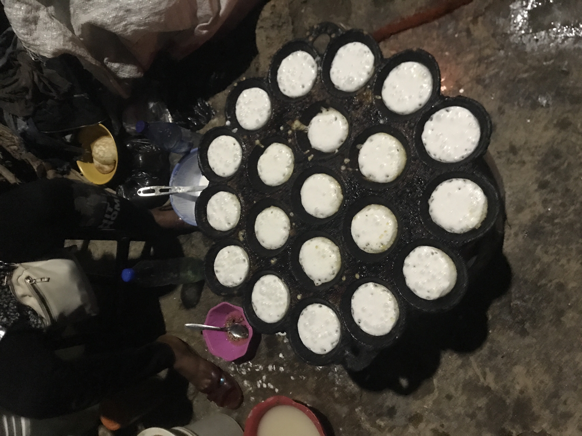 The image depicts an outdoor scene, likely a street food stall, in New Stadium, Nigeria. In the foreground, a dark, circular griddle with numerous small indentations holds several partially cooked, white, circular food items, possibly pancakes or dumplings. Some of the indentations are empty, indicating ongoing cooking. To the left, a red bowl contains a light-colored liquid, and a pink bowl with a spoon sits nearby. Behind these, a person's legs are visible, wearing dark trousers and a brown sandal. Further back, a white crossbody bag is slung over a shoulder, and a person in black clothing is partially visible. In the background, various items are piled up, including what appears to be fabric or blankets, a yellow bowl containing some food, and two plastic bottles, one blue and one clear. The ground is uneven and appears to be dirt or concrete, with some scattered white debris. The overall lighting suggests it is either evening or night, with artificial light sources not clearly visible but implied by the illuminated foreground.