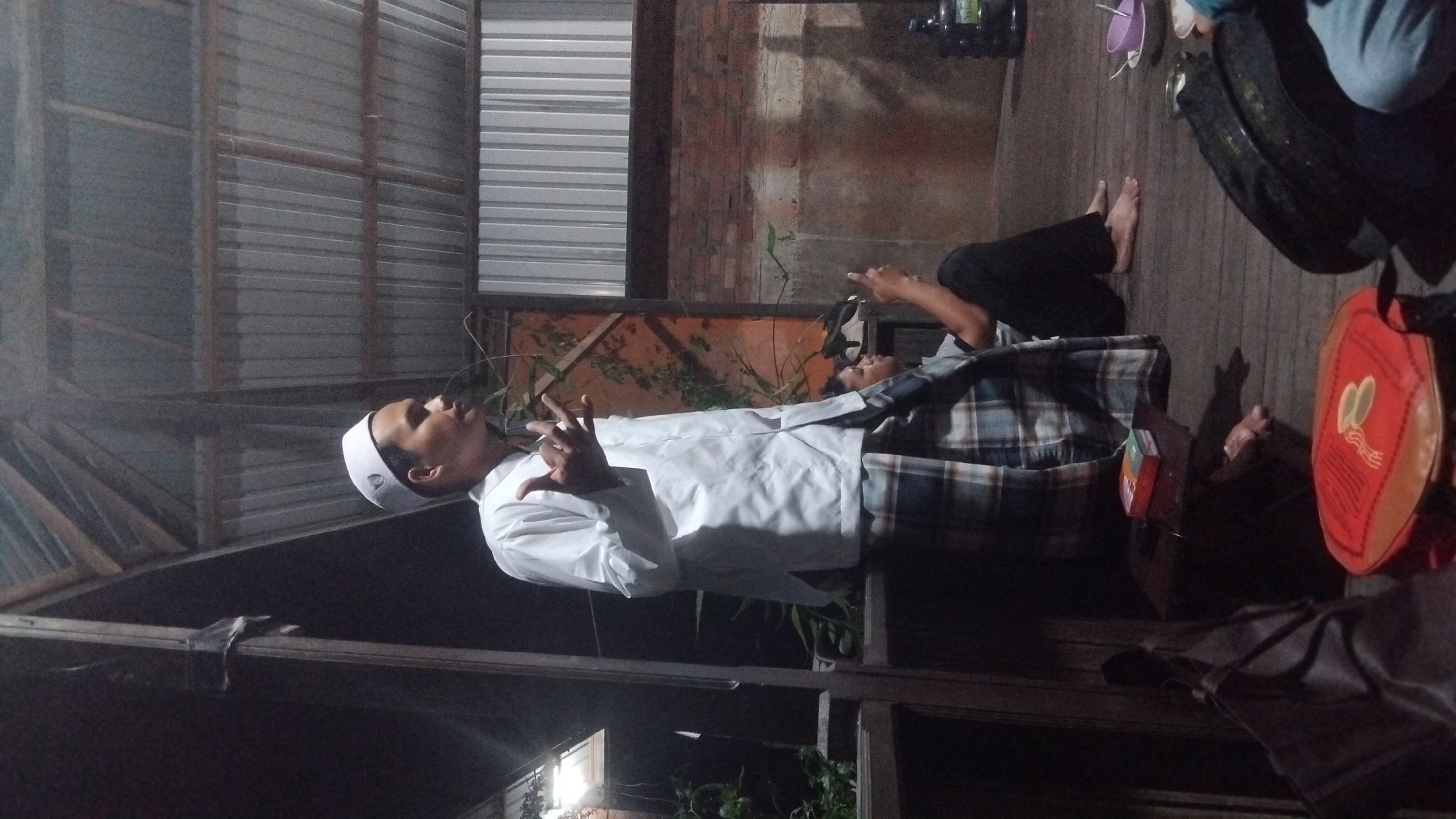 A man wearing a white peci and a white shirt with a plaid sarong stands on a wooden deck, gesturing with his hands. Another person sits on the deck to his right, looking at a phone. In the background, a corrugated metal roof is visible. The scene is dimly lit. The image appears to be taken in Bangun Rejo, Indonesia.