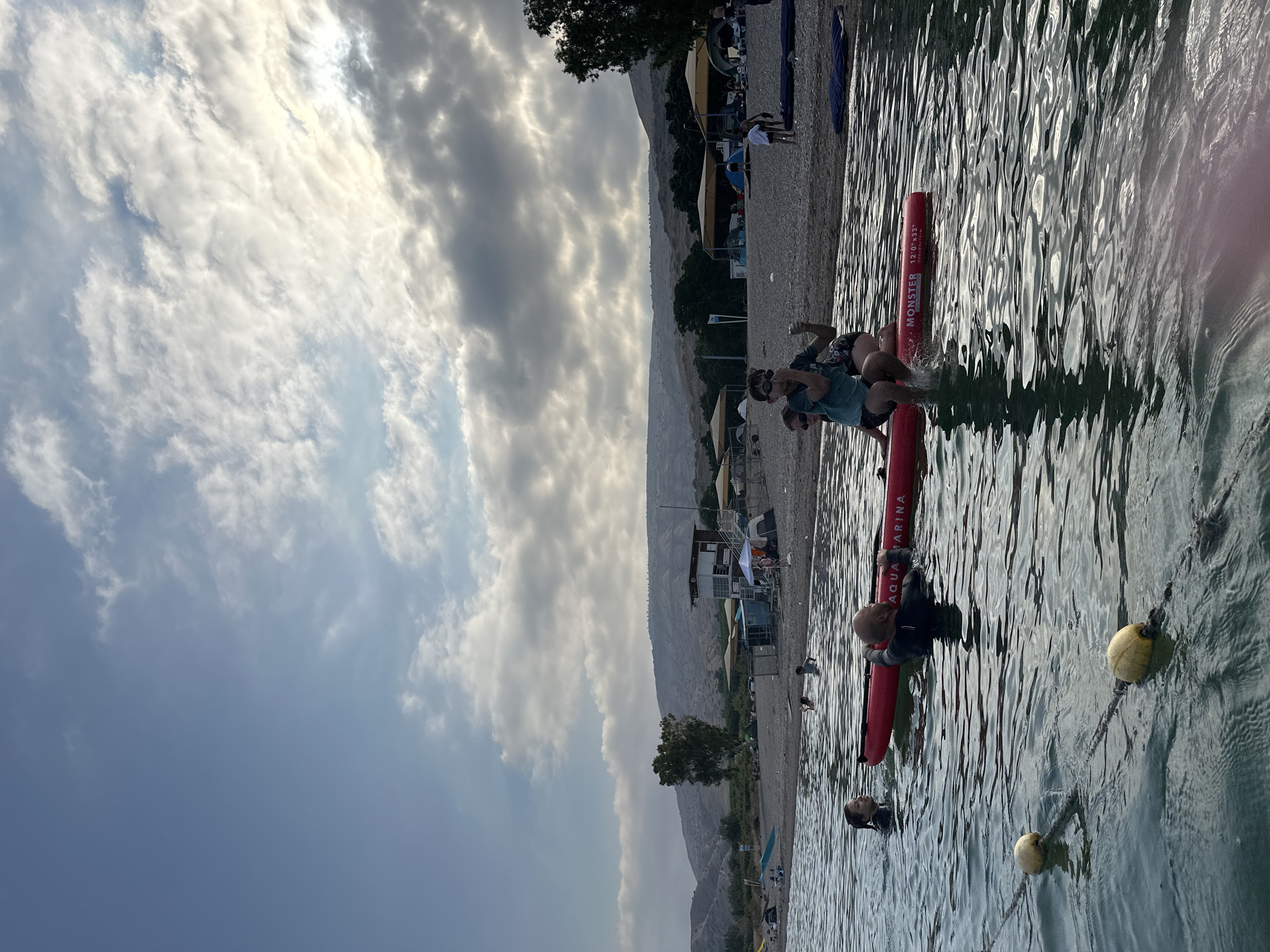 The image depicts a scenic view of the Sea of Galilee in Emek HaYarden Regional Council, Israel, under a partly cloudy sky. In the foreground, several people are in the water. A red inflatable stand-up paddleboard, branded "Aqua Marina Monster," is in the water, with two individuals sitting on it and one person partially submerged nearby. Further back, a beach is visible, lined with tents, umbrellas, and a lifeguard station. The landscape is characterized by rolling hills in the background. The overall atmosphere suggests a recreational day at the beach.