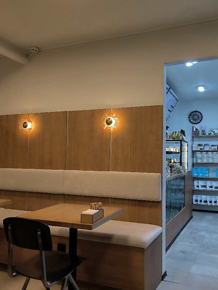 The image depicts the interior of a commercial establishment, specifically a cafe or coffee shop, located in Vinnytsia, Ukraine.
The foreground presents a seating area with walls painted off-white above and clad in vertical natural wood paneling below. Two circular wall sconces, featuring golden-orange textured diffusers, are mounted on the wood paneling and emit warm light. A long, light beige upholstered bench runs along the wall. In front of the bench, a square wooden table with a black metal pedestal base is positioned, accompanied by a single black chair with a metal frame. On the table, a small wooden caddy holds individually wrapped items, likely sugar packets or stirrers, and white napkins. The white ceiling displays a smoke detector and a segment of a pipe.
To the right, an open doorway or archway leads into a separate service area. This area is illuminated by cooler, bluish-white lighting. A menu board with dark background and white text is visible on the left wall, above a glass-fronted refrigerated display case. A large clock with Roman numerals hangs on the right wall. Below it, a shelving unit displays multiple organized packages in white and light blue, likely merchandise. A counter with a ribbed glass front runs along the immediate right side of the doorway. The floor in this background area is light-colored tile.
No individuals are present in the scene, and no actions or interactions are depicted. The environment appears clean and organized.
