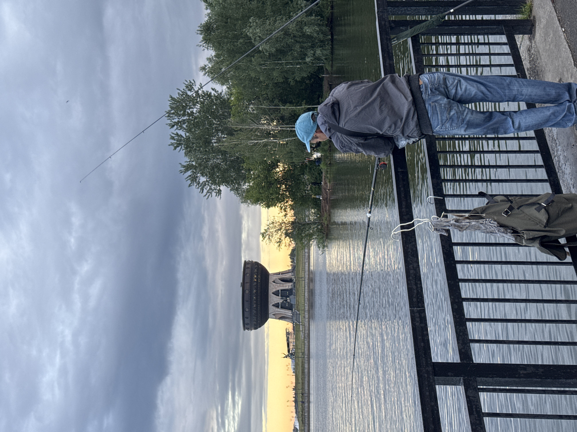 The image shows a man fishing off a bridge or pier. He is wearing a blue baseball cap, gray jacket, and blue jeans. He leans over the black metal railing, holding a fishing rod. Another fishing rod leans against the railing, and a green bag sits on the pier floor.
In the background, a body of water reflects the evening light. Beyond the water is the Qazan Family Center, a large, cauldron-shaped building, situated along the bank of the Kazanka River in Kazan, Russia. The sky above is overcast with a hint of sunset colors at the horizon. Trees are also visible in the background.