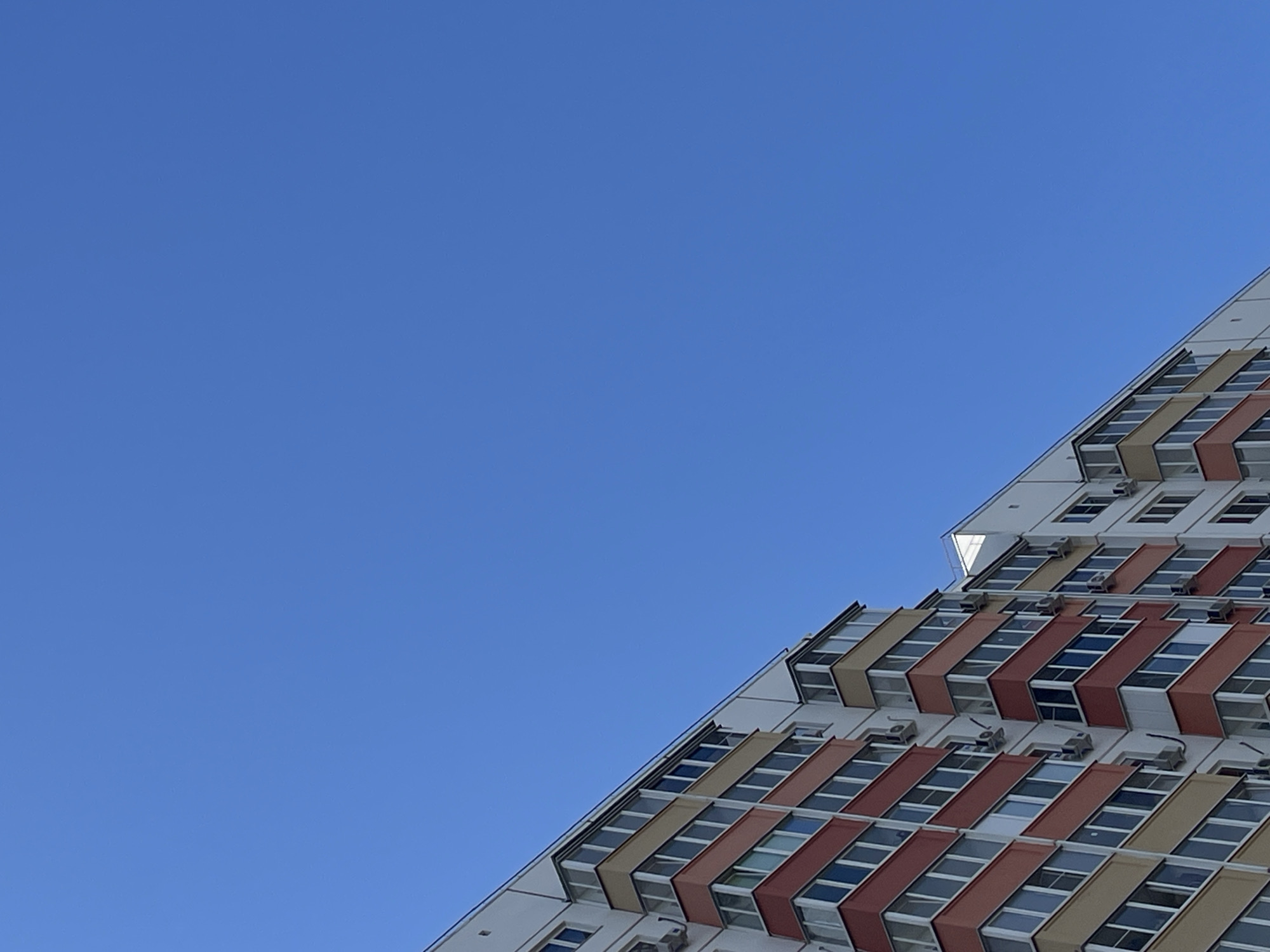A low-angle, full shot captures a section of a multi-story residential building against a clear, bright blue sky. The building features a facade with alternating horizontal bands of red and beige, interspersed with numerous windows and glass-enclosed balconies. Air conditioning units are visible mounted on the walls. The building's angled roofline is also apparent. The scene is devoid of people or any other discernible activity. The image is undated and its specific location within Rostov-on-Don, Russia, is not discernible from the visual information provided.