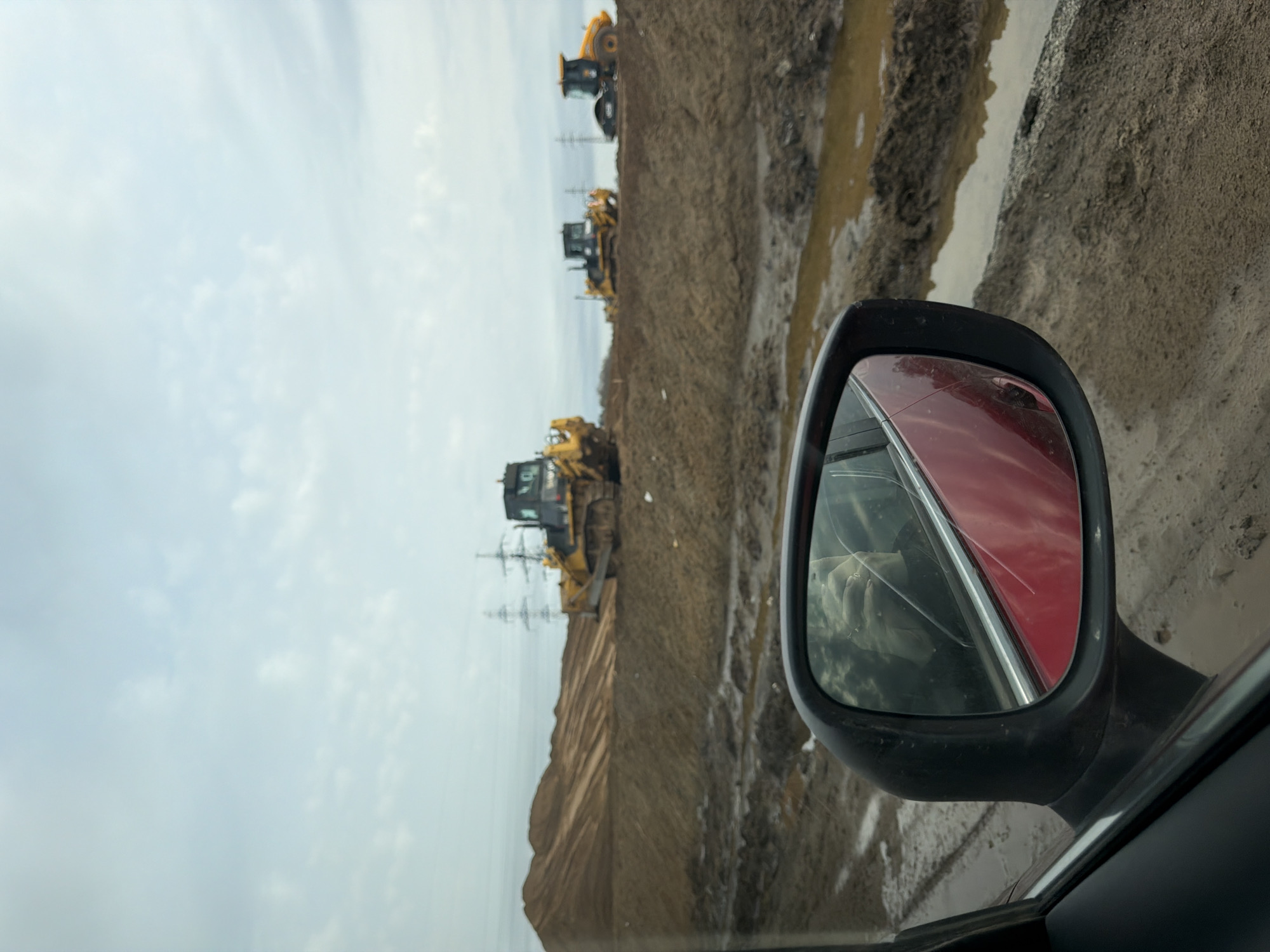 The image, likely taken from inside a car, shows a construction site near Zav'yalovo, Russia. In the foreground, the car's side mirror is visible, reflecting a red car. Beyond the car and the mirror, there is a muddy construction site with at least three bulldozers, one in the foreground and two others farther away. High-voltage power lines are visible near the bulldozers. The sky is overcast, and the overall scene suggests active construction or earthmoving work. Mud and disturbed earth are prominent, indicating recent activity at the site.
