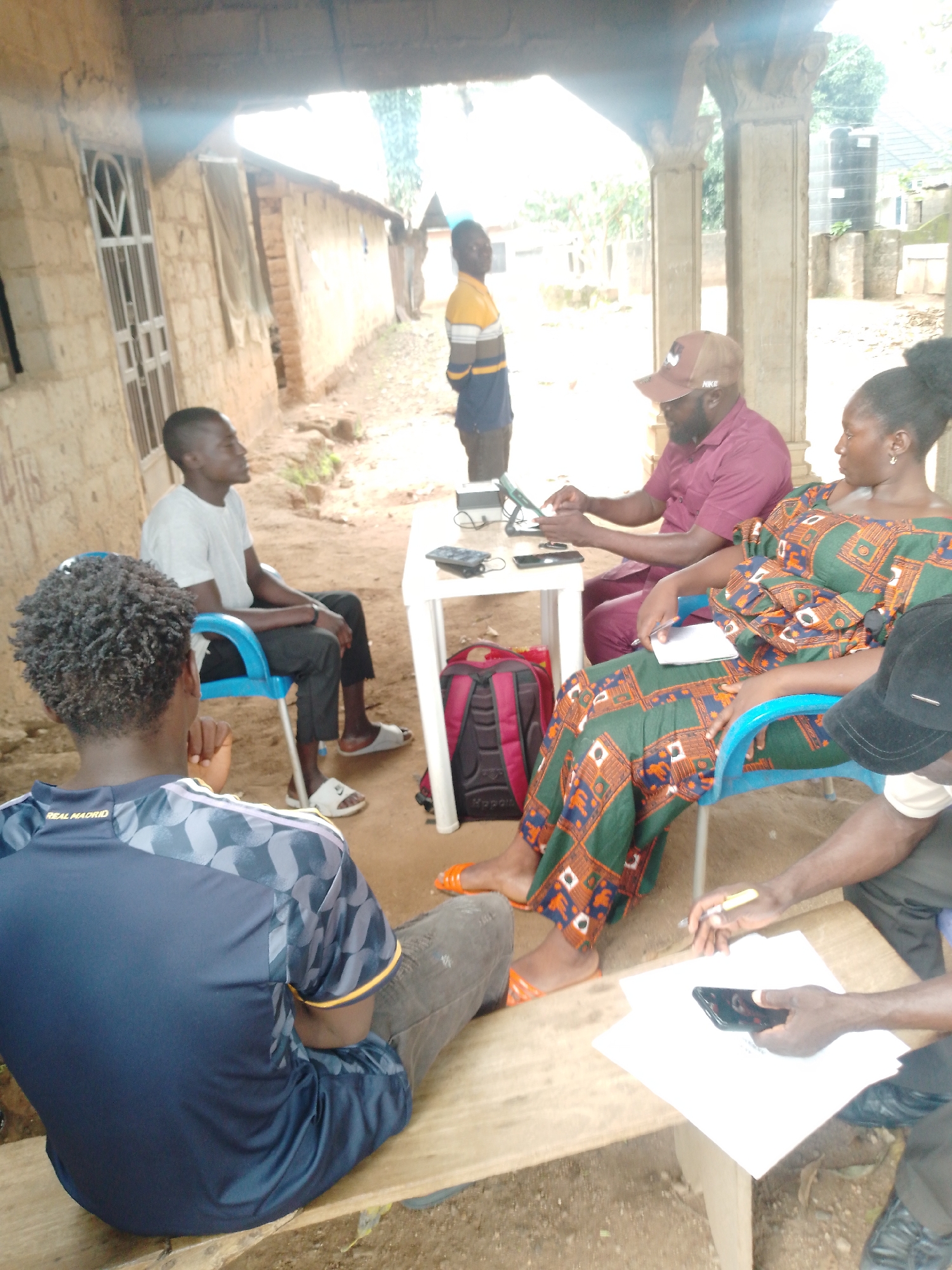 A group of six individuals, appearing to be adults, are gathered outdoors under a covered structure, likely a porch or shelter. The environment appears to be rural or semi-rural, with a building made of mud bricks or concrete blocks to the left and a water tank visible in the background. The ground is dirt and some debris.
Three individuals are seated in chairs around a white plastic table. One man, wearing a maroon polo shirt and a brown baseball cap, is seated and interacting with a tablet or laptop on the table. A woman, wearing a vibrant green and orange patterned dress, is seated next to him, holding a white notebook. Another man, wearing a white t-shirt and dark pants, is seated in a blue plastic chair to the left of the table, facing away from the camera.
Two individuals are seated on a wooden bench in the foreground, their backs to the camera. One is wearing a dark blue t-shirt with "REAL MADRID" visible on the back. The other is partially obscured.
A sixth individual, a young man wearing a yellow and blue striped shirt, is standing in the background, facing the building.
Objects on the table include the tablet/laptop, a smartphone, and possibly some papers. Two backpacks, one red and black, are on the ground near the table. Various personal items such as sandals and a pen are also visible. The lighting suggests it is daytime, possibly overcast.