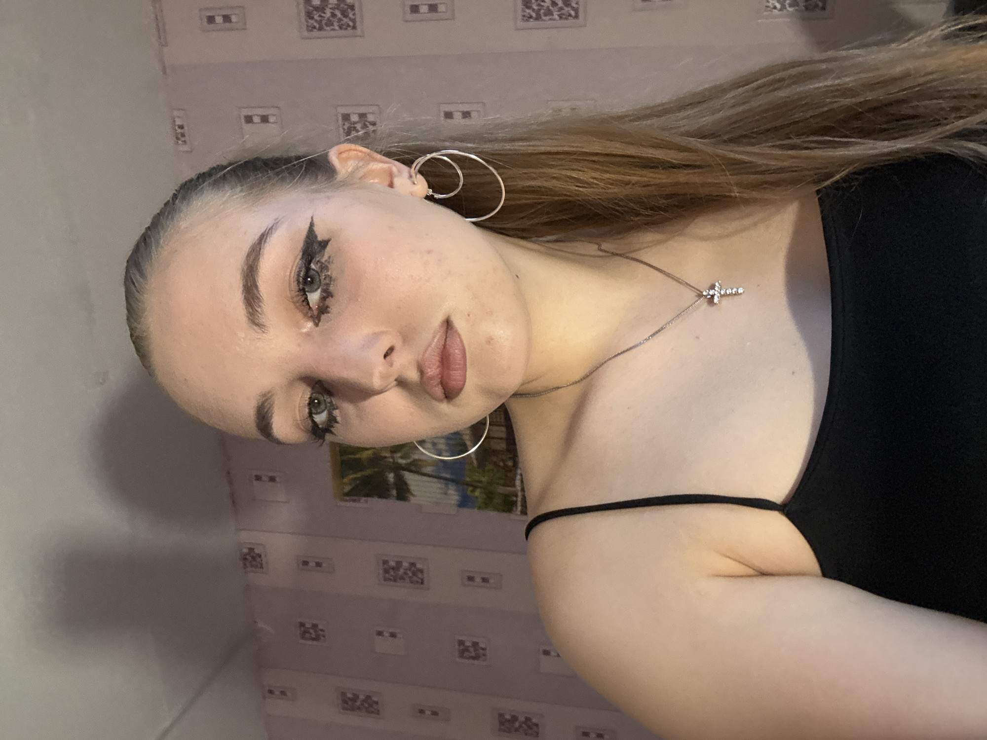 A young woman is posing for a portrait. She is wearing a black top, silver hoop earrings, and a necklace with a cross pendant. Her makeup includes dramatic eyeliner. The background features patterned wallpaper and a framed image. The image does not contain enough information to determine the location in Perm, Russia.