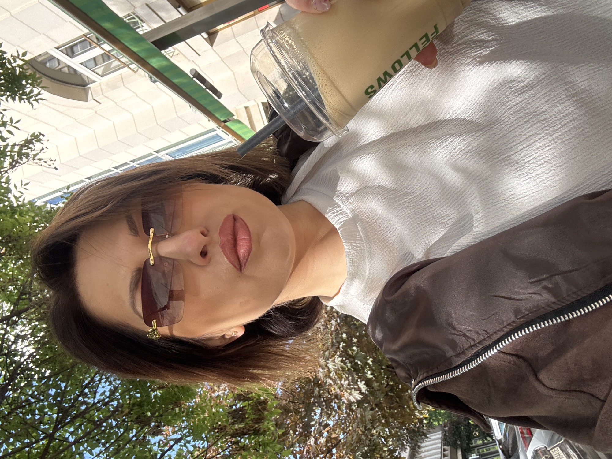 A woman in sunglasses and a white textured shirt, holding a coffee drink in a clear plastic cup, is photographed outdoors. The cup has the word "FELLOWS" printed on it. The woman also wears a brown bomber jacket. Buildings and trees are visible in the background. The location appears to be Sofia, Bulgaria, based on the architectural style of the buildings.