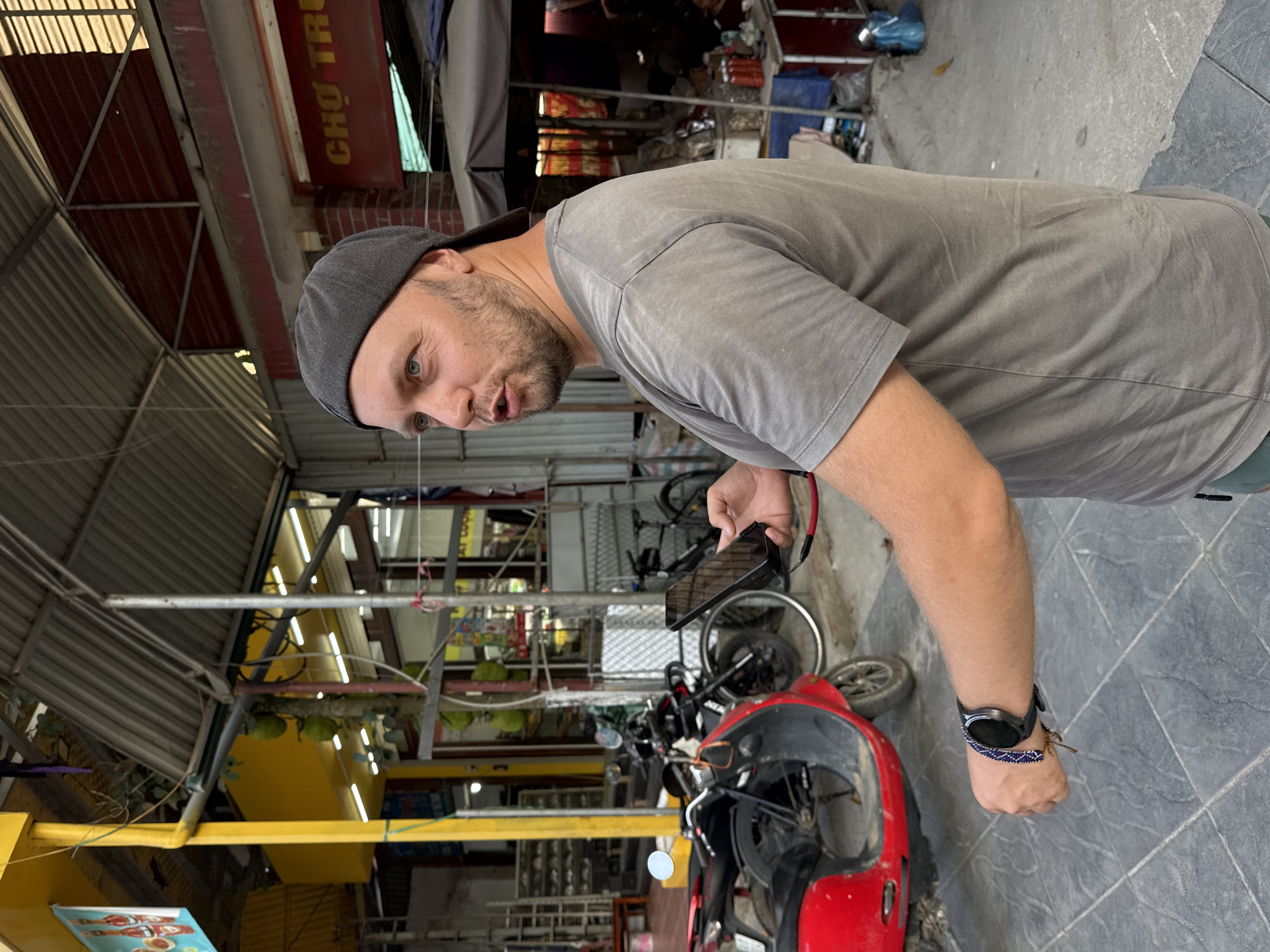 A mid-shot, rotated 90 degrees counter-clockwise, captures a Caucasian man standing in what appears to be a covered market or street in Phố Ràng, Vietnam. The man, with a short beard and light-colored eyes, wears a dark grey cap and a light grey short-sleeved t-shirt. He has a smartwatch with a blue and white patterned band on his left wrist and holds a black smartphone in his right hand. He looks towards the viewer with a slightly open mouth, suggesting he might be speaking or observing.
Behind him, a red scooter is parked, with other motorcycles or bicycles partially visible further back. The scene is characterized by a bustling market environment with various goods and stalls in the background. The ground is paved with grey tiles. Overhead structures consist of corrugated metal roofs, metal beams, and bright fluorescent strip lighting. Green gourds or fruits hang from an elevated fixture.
A red sign overhead prominently displays the Vietnamese text "CHỢ TRU", likely part of a market name such as "Chợ Trung". The overall impression is that of a busy daytime market in Southeast Asia.
