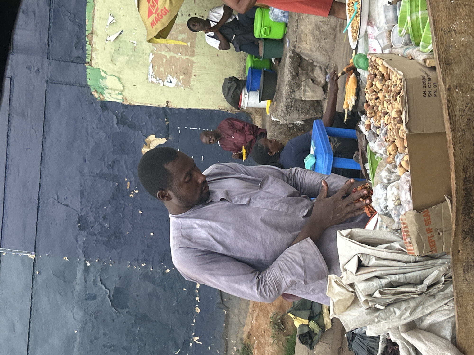 The scene depicts a street market in Jos, Nigeria. A man in a light grey robe sits near a stall selling roasted nuts and corn. He is handling some of the nuts. Behind him, other people are seated, including one wearing a red shirt and another wearing a head covering. The stall is stocked with roasted goods displayed in a cardboard box. Some items are also placed in bags. In the background, there's a building with walls that are painted in dark blue and yellow-green, with signs of age and wear. Other vendors and customers can be seen in the background, along with containers and various items for sale.