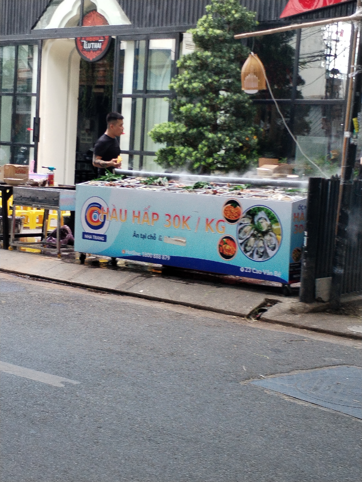 The image depicts a street scene in Bac Nha Trang Ward, Vietnam. In the foreground, a gray asphalt road with a white lane marker is visible. A blue and white food cart, adorned with images of seafood and text in Vietnamese, is positioned on the sidewalk. The cart displays the text "ỐC HẤP 30K/KG" and "NHATRANG," indicating it sells steamed snails for 30,000 Vietnamese Dong per kilogram and is located in Nha Trang. A man in a black t-shirt stands behind the cart, holding a piece of food. Behind the cart, a building with large windows and a prominent red sign with Vietnamese text is visible. Green trees and other vegetation are also present in the background. The overall scene suggests a bustling street-side food vendor.