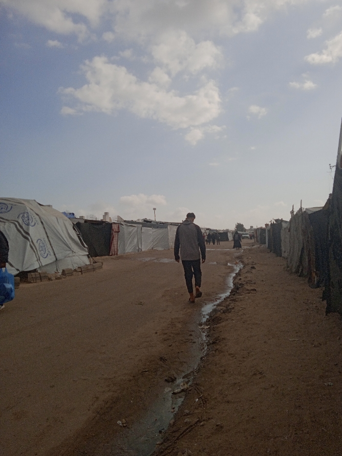 The image depicts an outdoor scene in Khan Yunis, Palestinian Territory, showing a dirt path flanked by rows of makeshift shelters beneath a sky with scattered white clouds. A male figure, viewed from behind, walks away from the camera along the path, wearing a grey long-sleeved top and dark trousers; his right foot appears bare or in open-toed footwear. A narrow channel of muddy water runs along the right side of the path. To the left, a large, light-colored tent with blue markings is visible, its base secured by logs. On both sides of the path, various improvised shelters, constructed from materials like tarpaulins, extend into the distance. In the background, several indistinct figures are visible further down the path, and taller structures rise above the low-lying shelters.