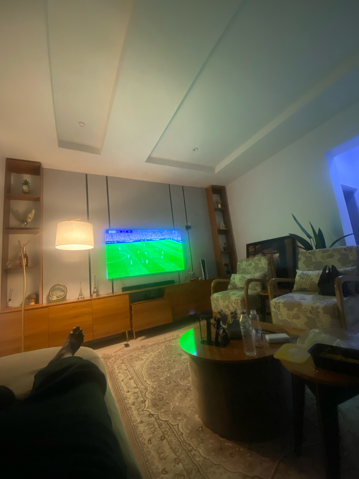 The image shows a living room scene with a television displaying a football match, a sofa with a person's legs visible, two armchairs, a coffee table, and a floor lamp. The room has a modern design with a dropped ceiling and wooden furniture. The overall lighting is dim, with the television screen and floor lamp providing the primary light sources. There are also decorative items on the shelves and coffee table, including small Eiffel Tower replicas and plants. The scene appears to be set in Jos, Nigeria, as indicated by the implied context.